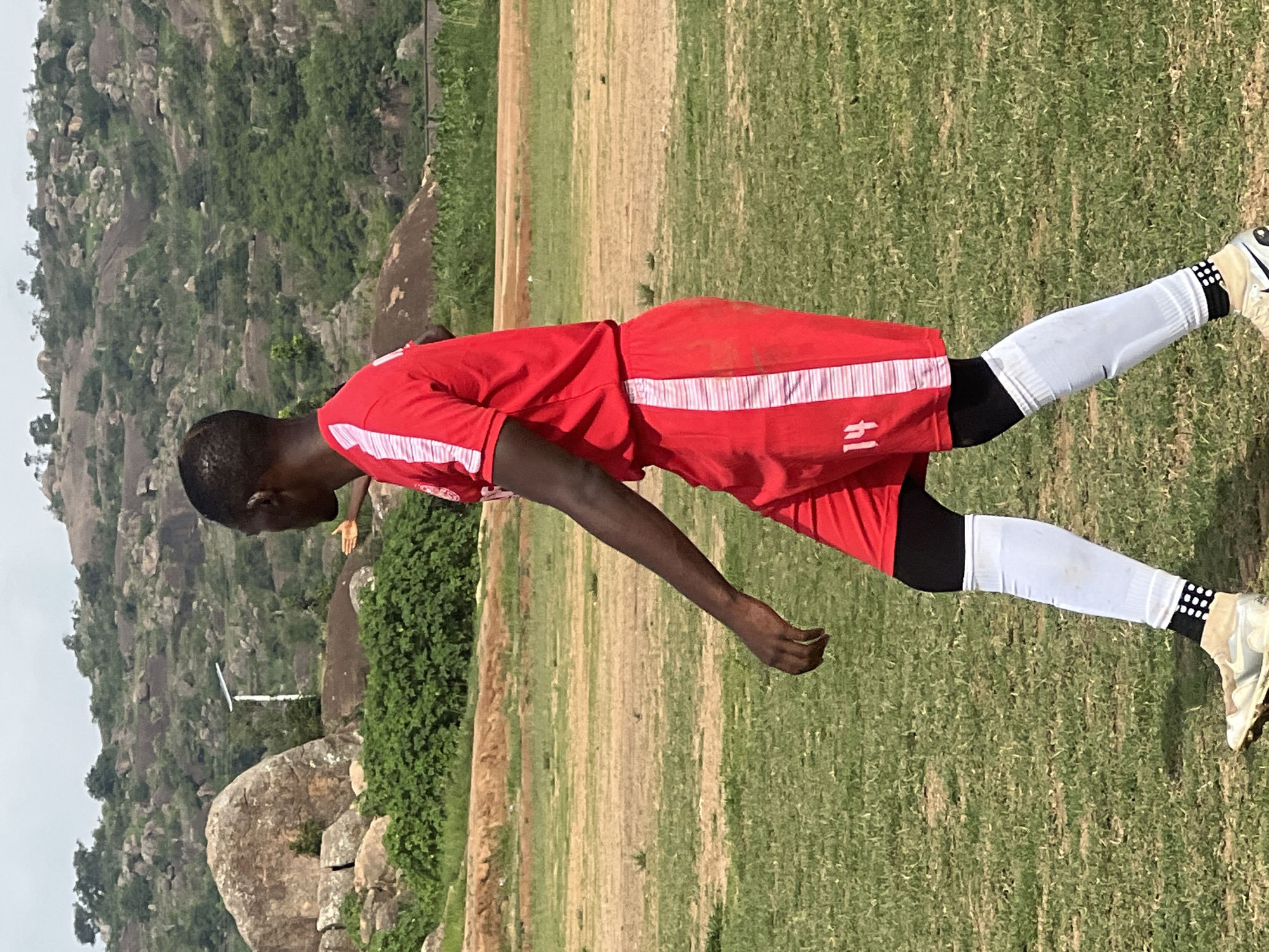 The image shows a young Black man in a red soccer uniform with white stripes down the sides of the shirt and shorts. He is wearing white socks with black accents at the top and bottom, and cleats. He is walking on a grassy field with a dirt path in the background. In the far background, there are rocky hills with sparse vegetation, characteristic of the landscape in Jos, Nigeria. The sky is overcast. The man appears to be in motion, with his left arm slightly extended and his right arm at his side. His number "14" is visible on his shorts.