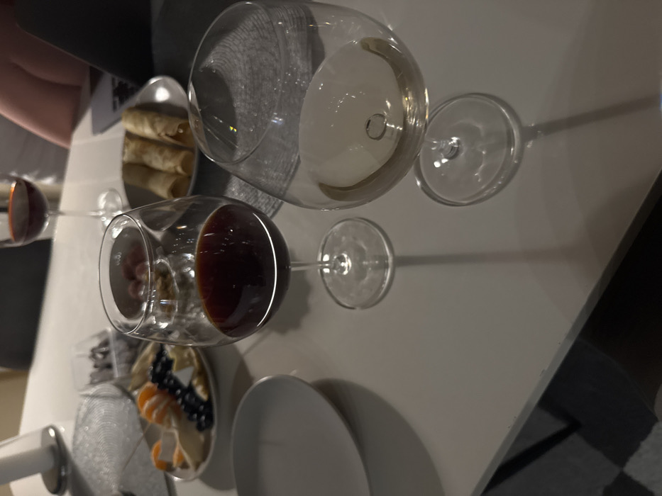 A table is set with a sophisticated spread, featuring two wine glasses filled with red and white wine. A plate of rolled crepes sits alongside a charcuterie board garnished with fruit and cheeses. A pristine white plate is also part of the setting. The scene is brightly lit, casting long shadows of the glasses on the table.