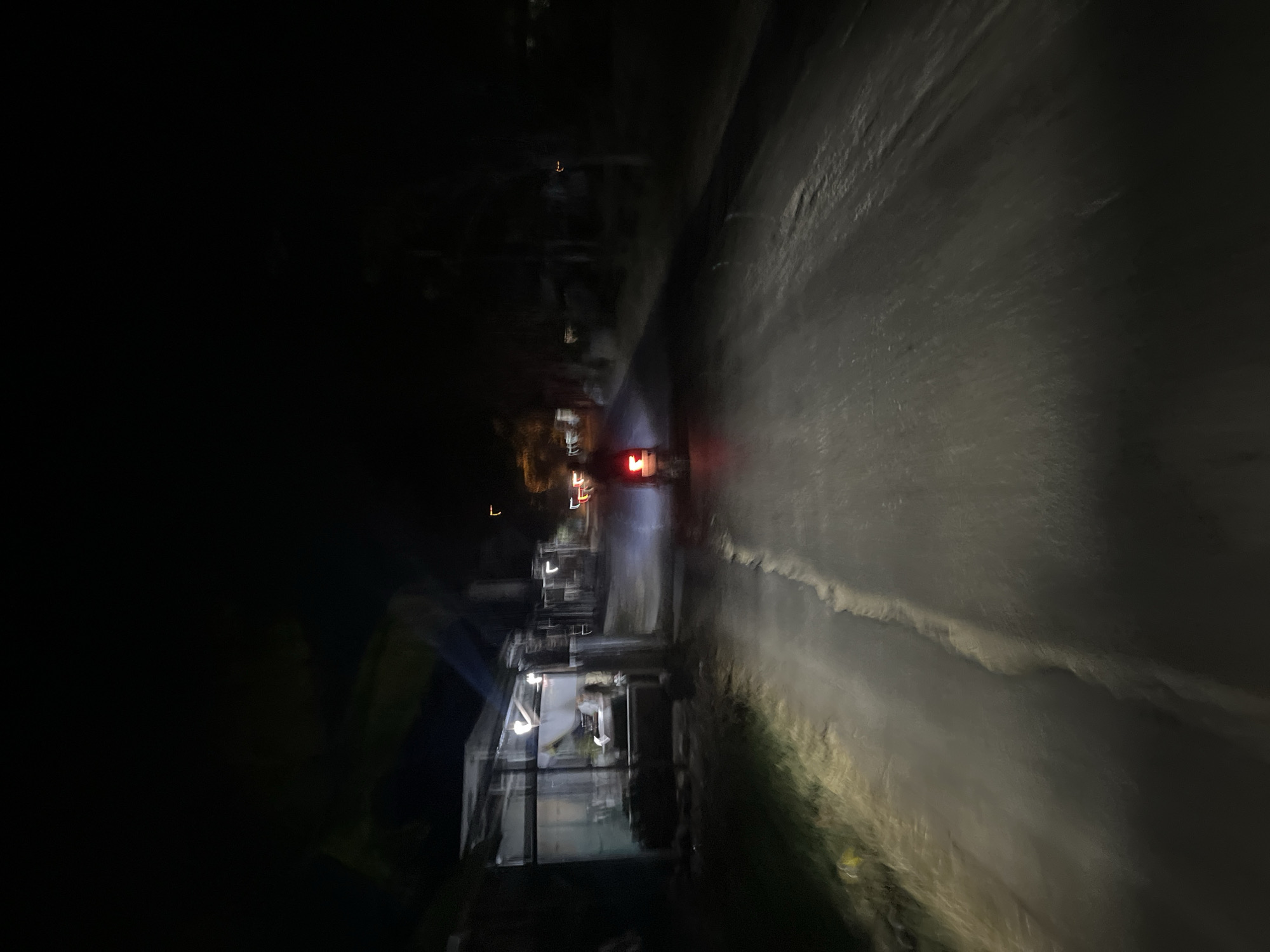 The image captures a dimly lit street scene at night in Pecangaan, Indonesia. A motorcycle with its taillights illuminated is the central subject, traveling away from the viewer down a paved road. The road is bordered by vegetation on the right and what appears to be a row of structures, possibly shops or houses, on the left. These structures are partially illuminated by artificial lighting, casting a cool blueish hue. The overall atmosphere is one of a quiet, rural or semi-urban street at night, with the primary light sources being the vehicle's lights and the interior lighting of the buildings. The image appears to have been taken from another moving vehicle, indicated by the motion blur.