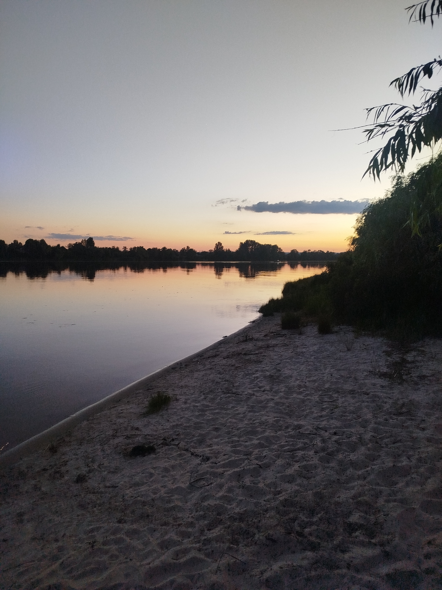 The image depicts a tranquil riverside scene at dusk. The sky exhibits a gradient of soft grays and pale oranges, with a few wispy clouds. A silhouetted treeline borders the opposite bank of the wide, calm river, which reflects the sky and the distant trees. In the foreground, a sandy shore slopes down to the water's edge. To the right, dense, dark green foliage, possibly willow branches, overhangs the scene, casting shadows on the lighter sand. The overall atmosphere is peaceful and serene. There are no people or animals visible in the image.