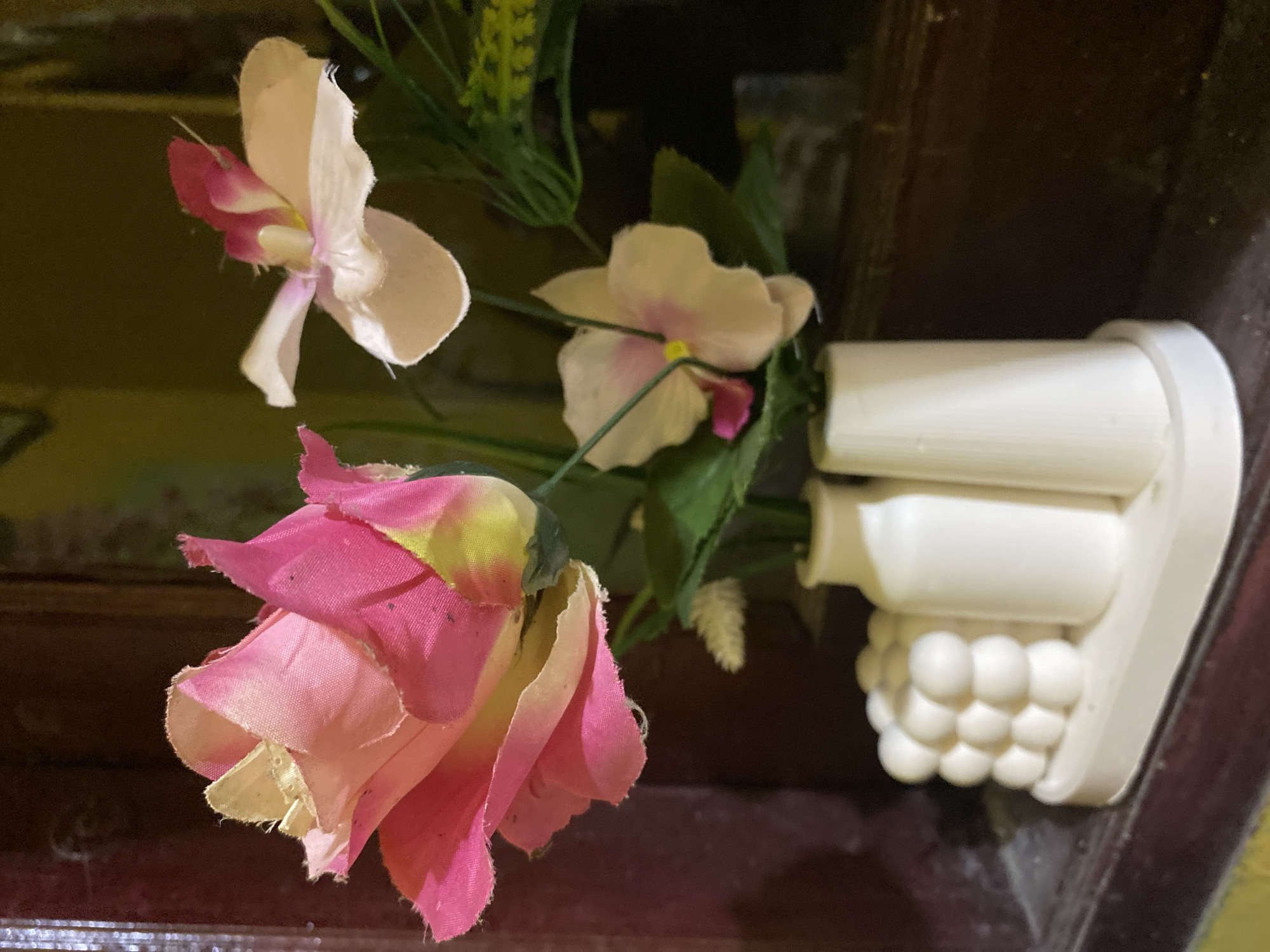 The image shows a floral arrangement in a white ceramic vase. The flowers include a pink rose, a white and pink orchid-like flower, and some green foliage. The arrangement is placed on a dark surface, possibly a table or shelf, with a blurred background suggesting an indoor setting. The location in Tanjongkapai, Indonesia, is not evident from the visual content.