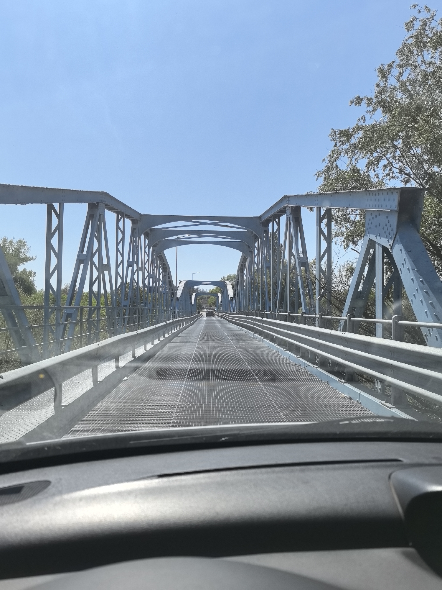 The image shows the view from inside a car driving across a blue metal bridge. The bridge has a steel grid deck and is supported by metal trusses. The sky is blue and clear. Trees are visible on either side of the bridge. The dashboard of the car is visible in the foreground. The location is the bridge over the Maritsa River in Simeonovgrad, Bulgaria.