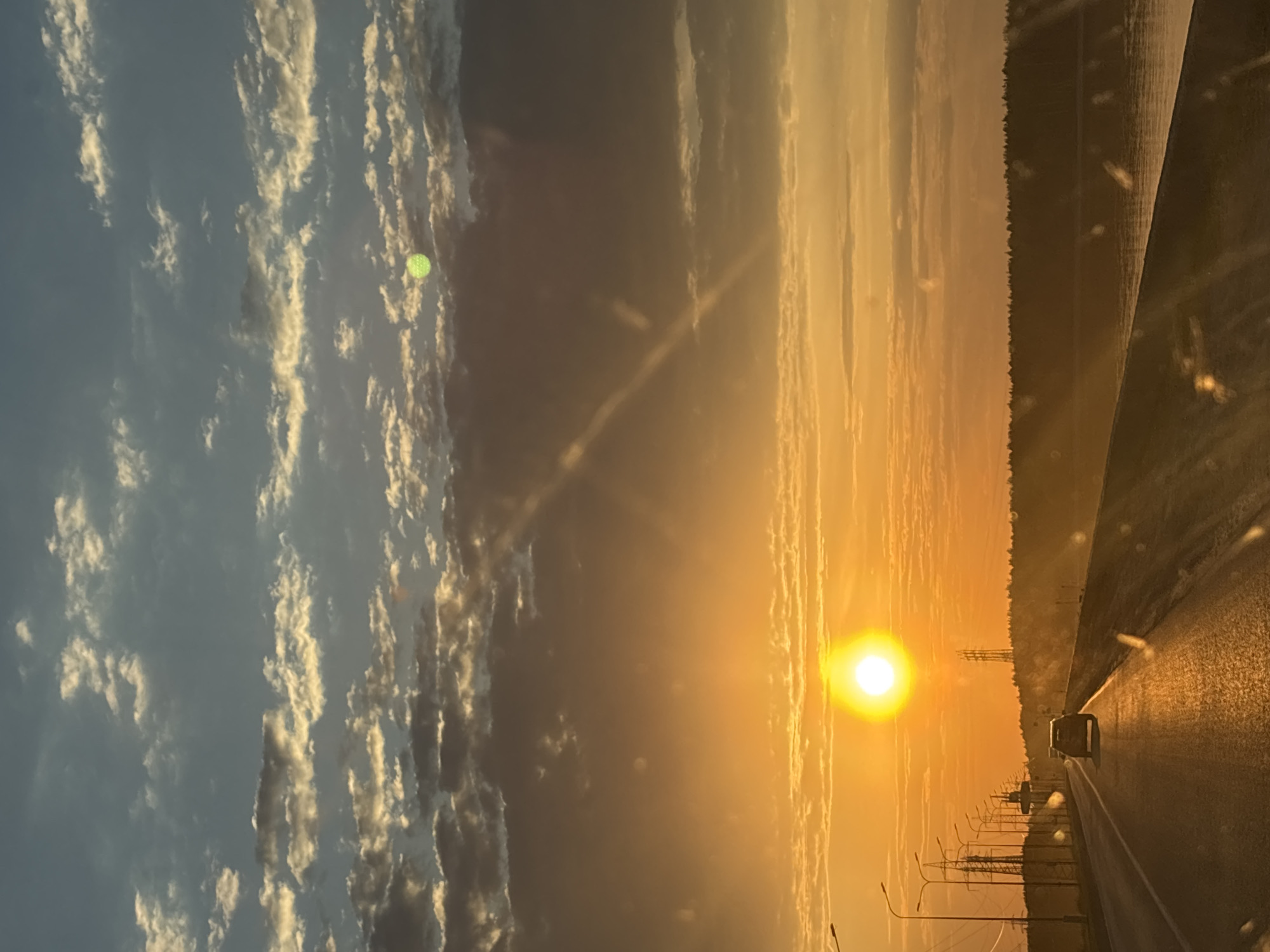 The image shows a sunset over a road in what appears to be a city outskirts, potentially near Chaikovsky, Russia. The sun is low in the sky, casting a golden glow across the landscape. To the left, clouds are visible in a partially blue sky. To the right, a highway stretches into the distance with a vehicle traveling on it. Streetlights and power lines are alongside the road. A dark forested area is visible on the right.