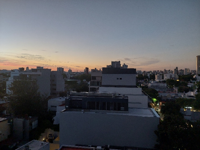 An urban landscape unfolds under a dusky sky, tinged with the last light of sunset. The cityscape is densely packed with buildings of varying heights and styles. The foreground is dominated by rooftops and structures, leading towards a more distant skyline dotted with high-rises. Patches of green suggest trees interspersed among the buildings. Streetlights hint at the onset of evening in the city. The image is primarily composed of cool gray and blue tones, with warmer hues emanating from the horizon.