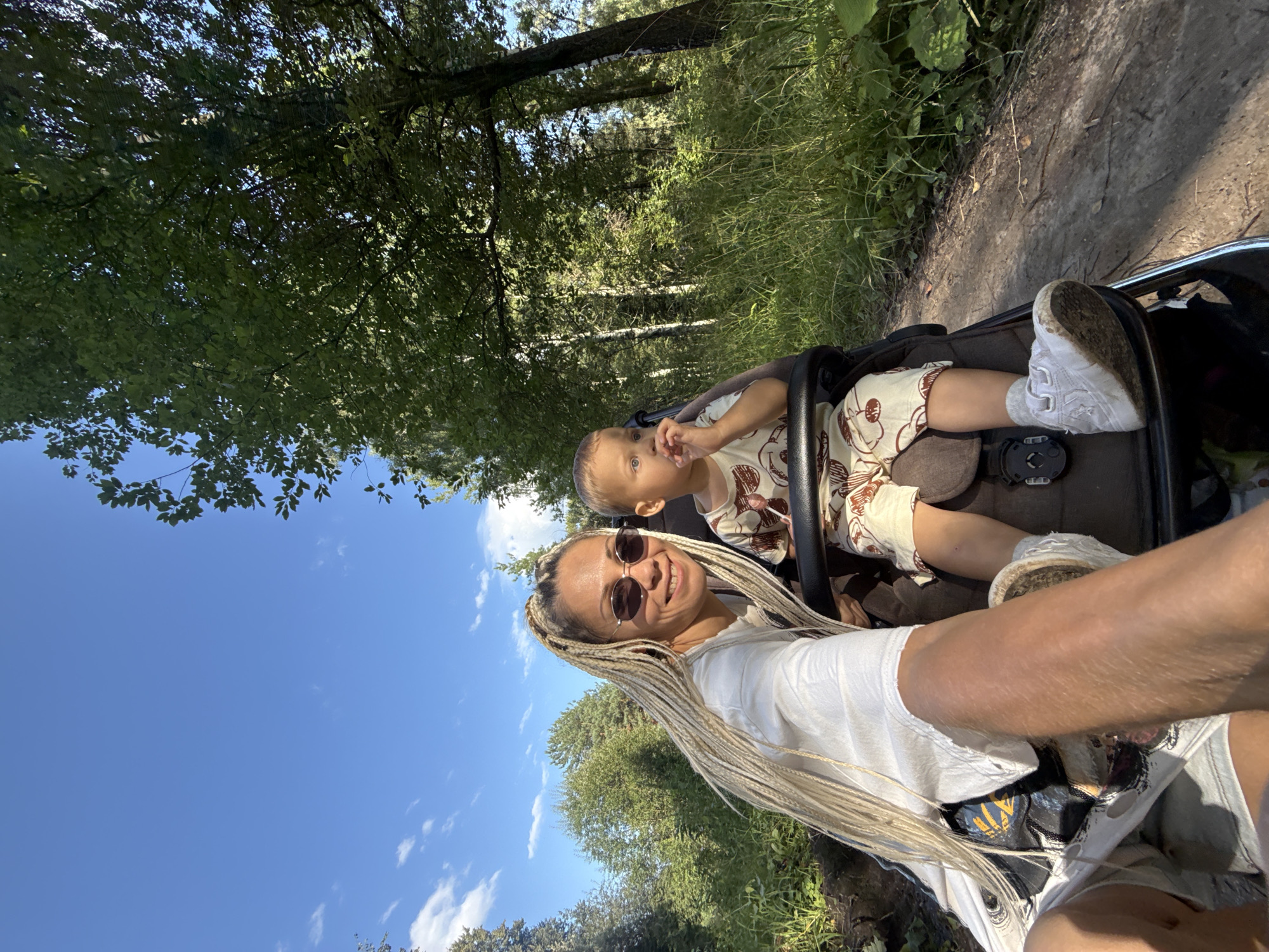 A woman with blonde braided hair and sunglasses is taking a selfie in a stroller in a wooded area. A young child is seated next to her in the stroller, looking to the side and chewing on something. The woman is wearing a white t-shirt and shorts, while the child is in a patterned outfit. They are on a dirt path, with lush green trees and foliage surrounding them. The sky is blue with some scattered clouds. The scene appears to be a park or natural area, possibly in or near Rusanovo, Russia, given the visual context of birch trees common in that region.