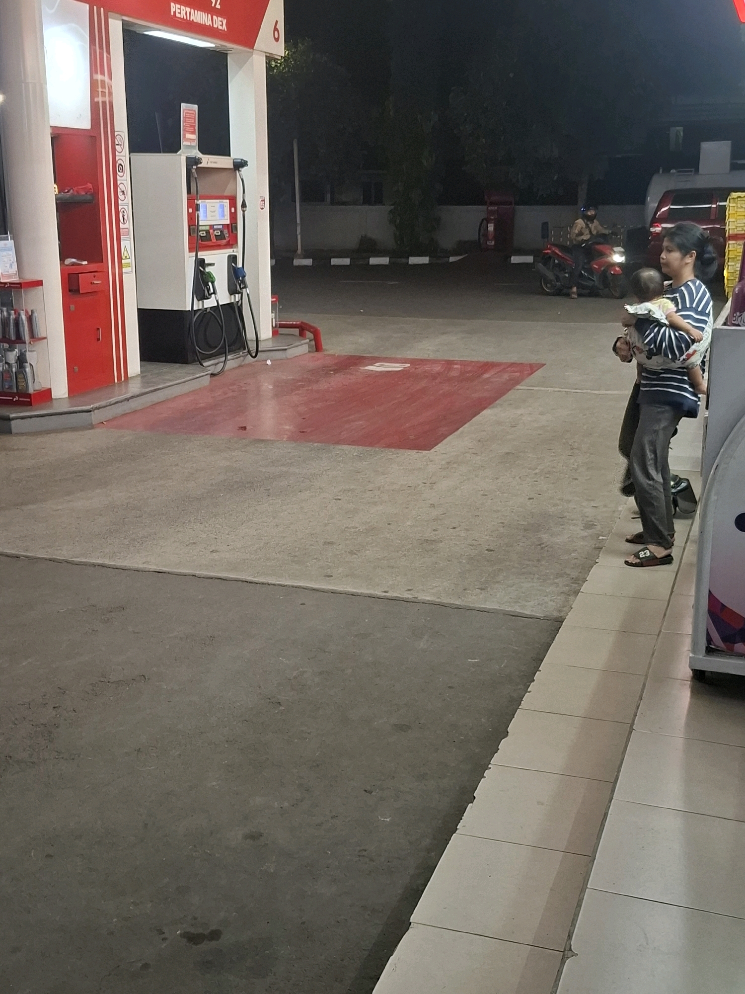 The image depicts a nighttime scene at a Pertamina Dex gas station in Karangtengah, Indonesia. A woman is holding a baby while standing near a gas pump. To the left, the gas pump station is visible, with hoses and a payment terminal. In the background, a person is riding a motorcycle. The ground is paved concrete, with a red-colored area in front of the gas pumps. The overall lighting is dim, characteristic of an outdoor nighttime environment.