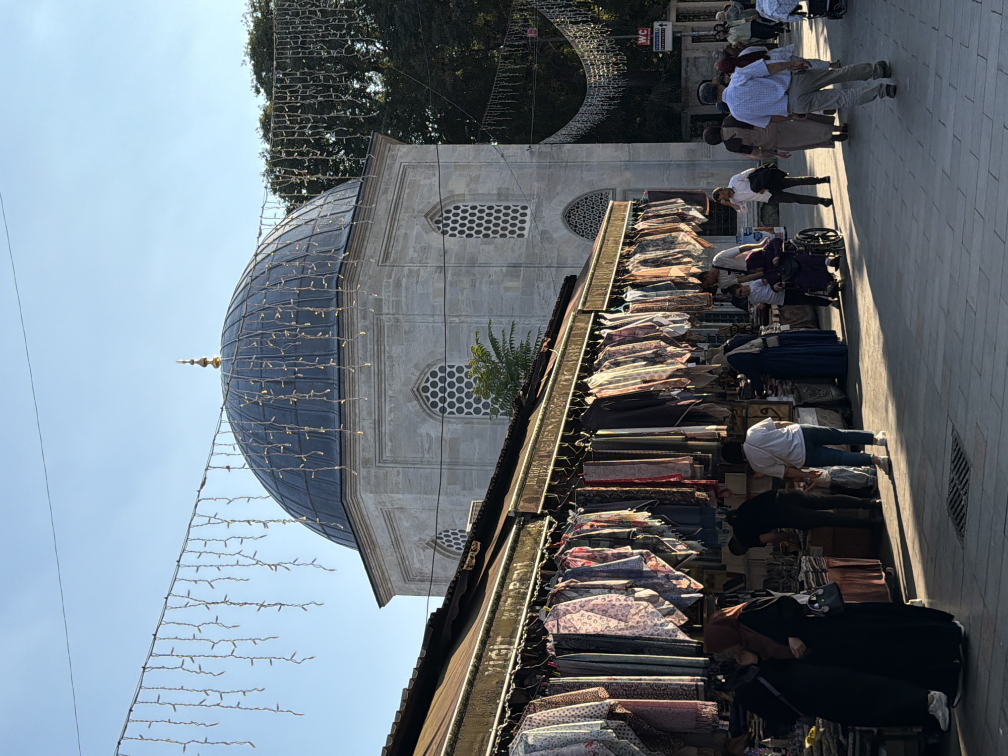 In Eyüpsultan, Turkey, an outdoor market thrives under a bright, clear sky. The market stalls, adorned with strings of decorative lights, overflow with a colorful display of fabrics, scarves, and textiles. Shoppers, dressed in a mix of traditional and modern attire, navigate the paved walkway, some browsing the merchandise, others engaged in conversation. A prominent domed structure, likely a mosque, stands as a backdrop to the bustling scene. The architectural style of the building, with its arched windows and intricate stonework, suggests historical significance. The atmosphere is lively, with a sense of community and commerce intermingling.