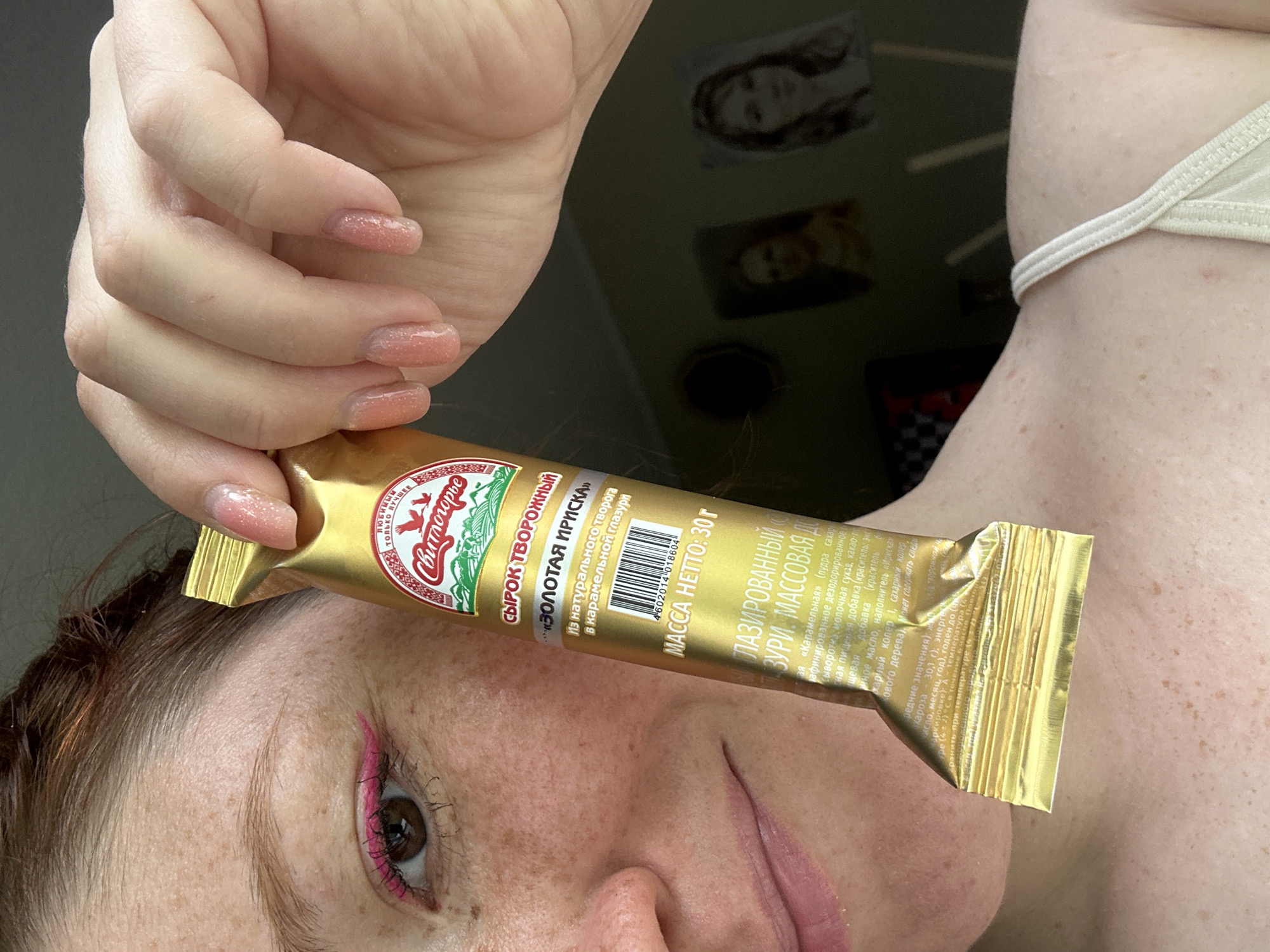The image shows a person holding a golden-colored package of "Svitlogorye" brand "Golden Iriska" glazed curd snack. The person has fair skin, freckles, and auburn hair. They are wearing pink eyeliner. The package is labeled in Russian, indicating it is a curd snack with a caramel glaze. The text on the package includes the product name, weight (30g), ingredients, and nutritional information. The background is blurry and shows what appears to be personal photos on a surface, and a hint of a white article of clothing, possibly a bra strap, near their neck. The scene appears to be indoors. There's no indication of Nizhny Novgorod or Russia in this frame aside from the product being labeled in Russian.