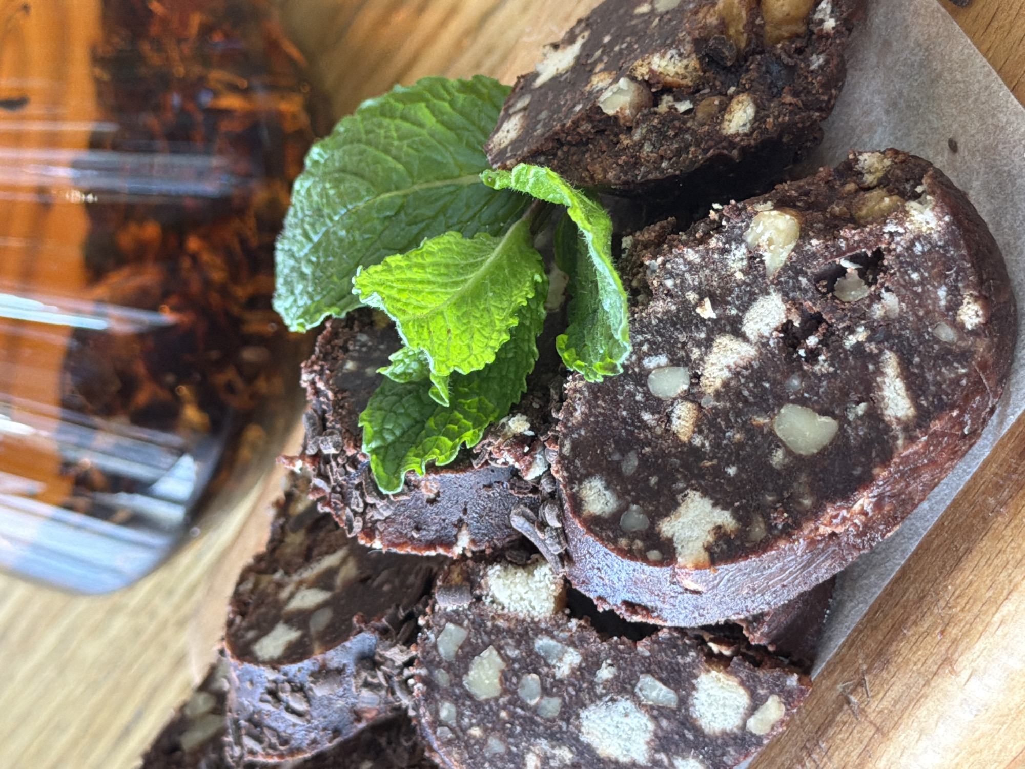 The image captures a close-up, slightly overhead view of sliced chocolate salami, or chocolate sausage, artfully arranged on a wooden surface. Several circular slices of the dark brown confectionery are visible, showcasing a rich texture studded with irregular, light-colored pieces, likely biscuits or cookies, and lighter brown chunks of nuts, possibly walnuts or pecans. The edges of some slices appear slightly crumbly.
A cluster of fresh, vibrant green mint leaves serves as a garnish, prominently placed on top of some of the chocolate slices, adding a pop of color and a hint of freshness to the rich dessert. The chocolate salami is presented on a piece of off-white paper, possibly parchment paper, which rests on a light-colored wooden cutting board or table, showing distinct grain.
In the blurred background on the left, a transparent glass jar or bottle is partially visible. It contains a dark, textured substance immersed in a liquid, which could be an infused beverage, dried fruits, or tea.
The scene suggests an indoor setting, possibly a kitchen or cafe, with bright, natural lighting highlighting the mint leaves and reflecting off the glass jar. The overall impression is one of a prepared dessert or snack ready for enjoyment. There are no people visible, and no discernible text. While the location context is Krasnoyarsk, Russia, there are no specific visual cues within the image itself to confirm this geographic detail.