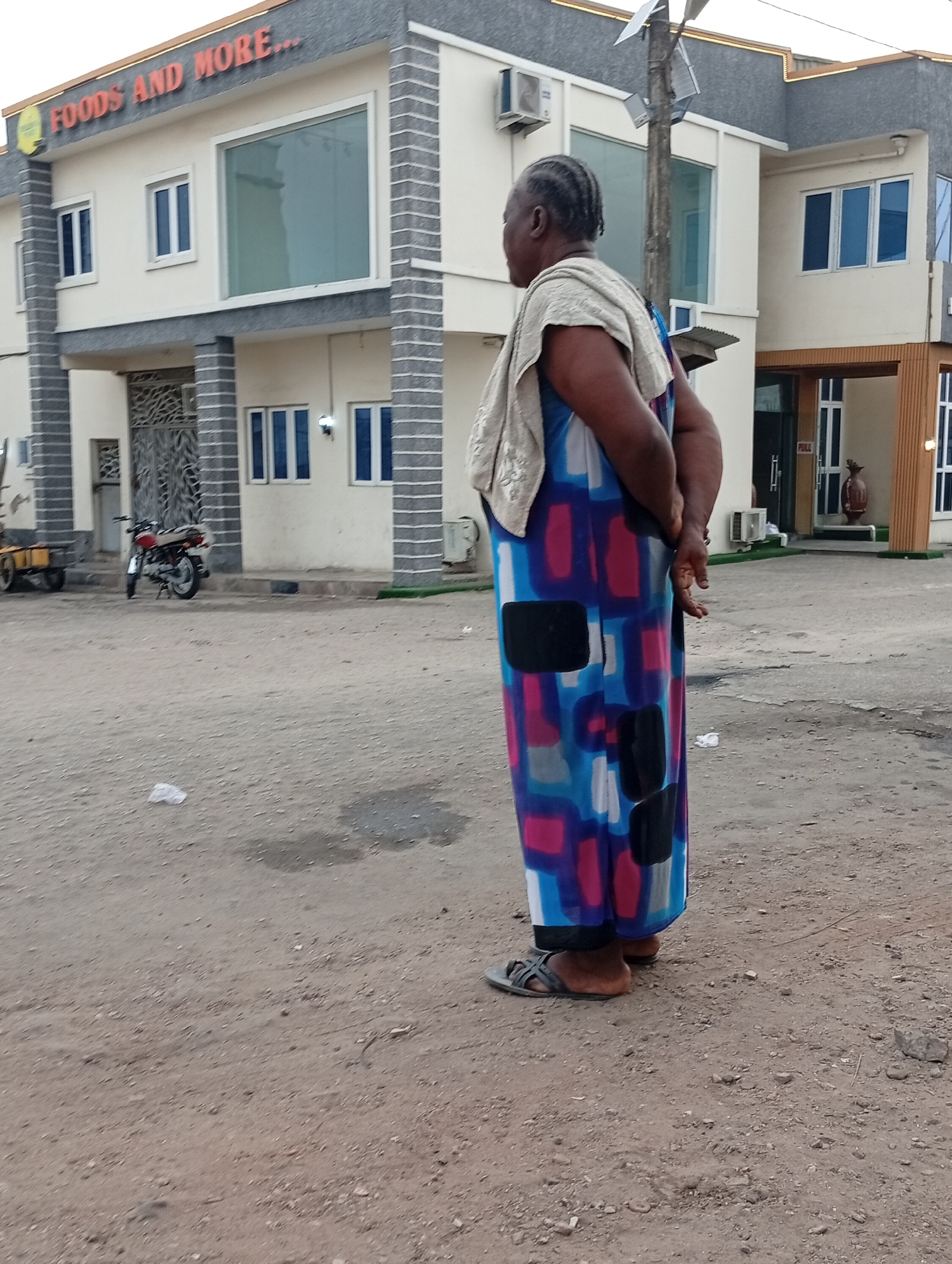 A woman stands with her back to the camera in front of a building labeled "FOODS AND MORE..." in Lokoja, Nigeria. She wears a colorful dress with blue, red, and black rectangular patterns and gray sandals. A cream-colored cloth is draped over her shoulders, and her hands are clasped behind her. The building is light beige with gray trim and has multiple windows. A motorcycle and a small yellow cart are parked to the left of the building entrance. The ground is dirt.