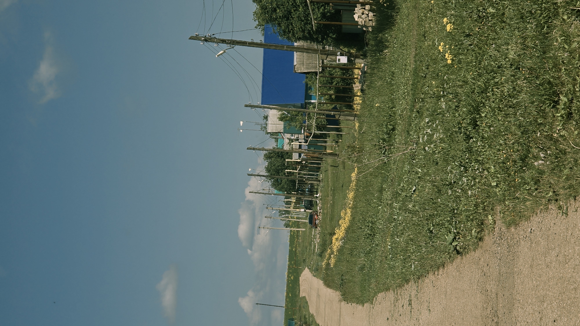 The image depicts a rural scene with a dirt road curving into the distance on the left. The road is bordered by a grassy field with scattered yellow and white wildflowers. In the background, a row of houses with blue and green roofs can be seen, interspersed with trees and utility poles carrying electrical wires. A few cars are parked along the road. The sky is blue with a few scattered clouds. The location is identified as Kzyl-Chishma, Russia.