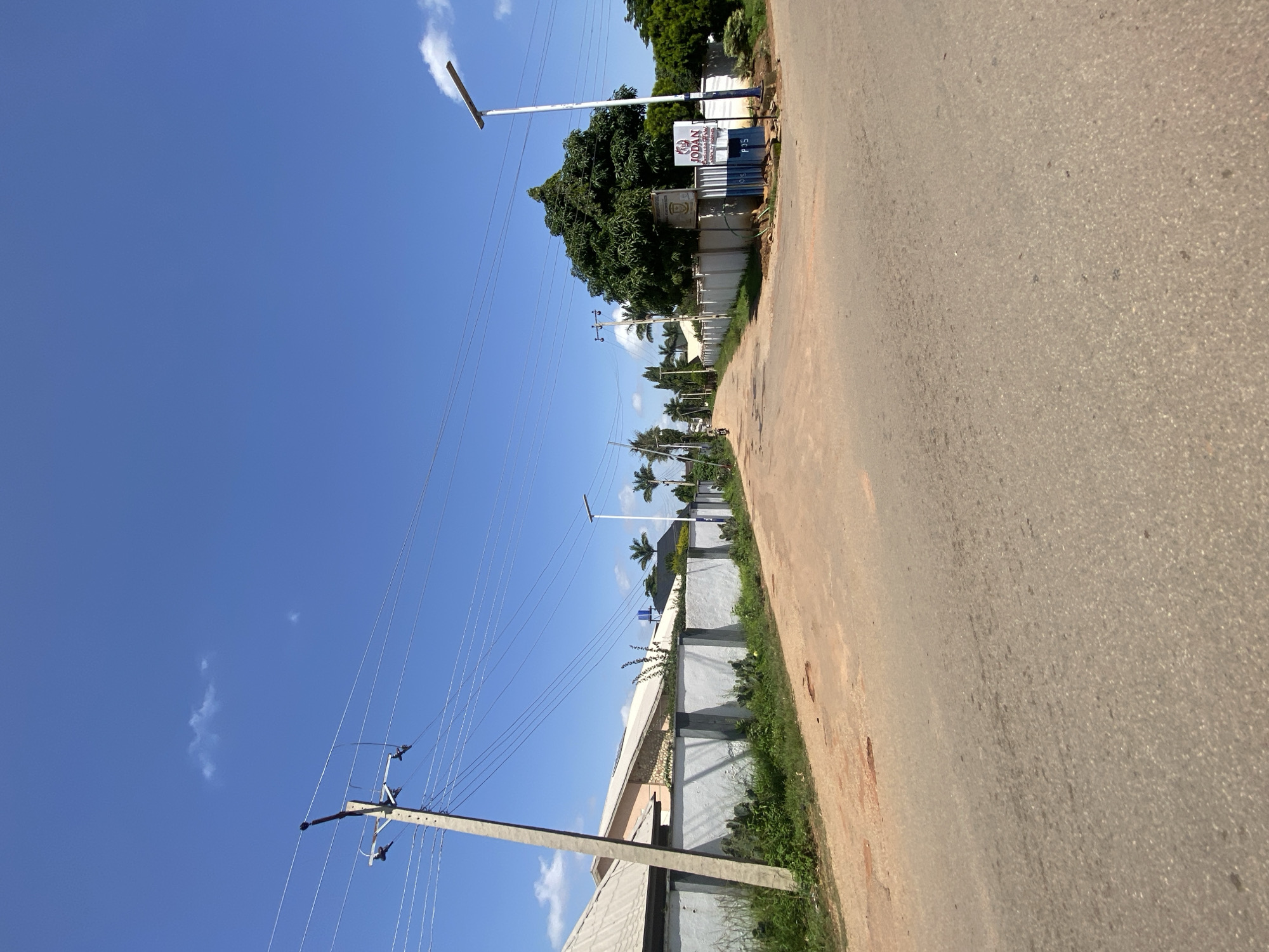 The image depicts a street scene in Gura Top, Nigeria. A paved road with some patches of dirt leads into the distance. On the left side of the road, a concrete wall with dark gray pillars borders properties with houses featuring tiled roofs and some stone accents. A prominent utility pole with wires stands to the left. On the right side of the road, another concrete wall is visible, with a large tree and a sign indicating "JODAN" and "POST 015". A modern street lamp is positioned near the sign. The sky is clear and blue with a few small white clouds. Palm trees and other greenery are scattered throughout the scene. A motorcycle is visible in the distance on the left side of the road. The environment appears to be a residential or commercial area within the city.