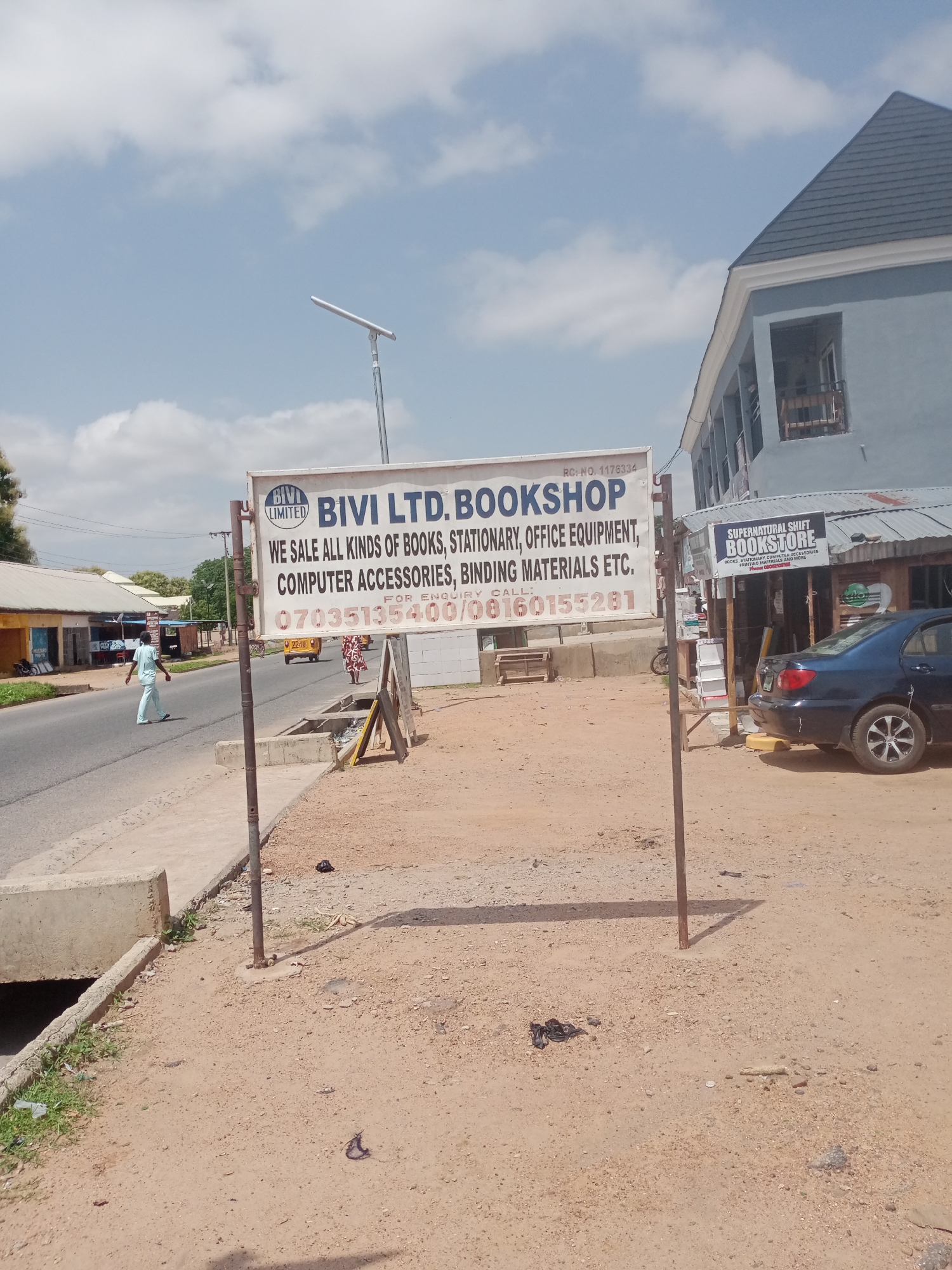This media file captures a street scene in Jalingo, Nigeria, likely during the daytime under a partly cloudy sky. The focal point is a large, white signboard for "BIVI LTD. BOOKSHOP." The sign states they sell "ALL KINDS OF BOOKS, STATIONARY, OFFICE EQUIPMENT, COMPUTER ACCESSORIES, BINDING MATERIALS ETC." Contact numbers are prominently displayed. To the right of this sign, a smaller sign for "SUPERNATURAL SHIFT BOOKSTORE" is visible, advertising similar items.
In the background, a light blue, two-story building with a dark roof is visible. A dark-colored sedan is parked in front of the bookstore. On the road, a yellow vehicle and several pedestrians are present. One person, dressed in light blue attire, is walking across the street. The environment appears to be a commercial area with buildings lining the road and a paved sidewalk. The ground is sandy and dusty, typical of many Nigerian landscapes. The overall impression is that of a bustling roadside in a Nigerian town.