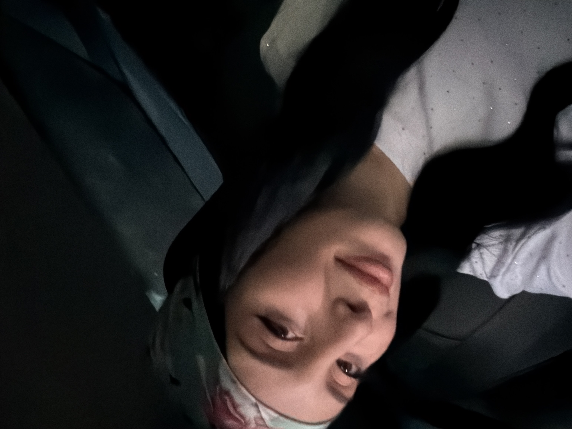 A medium shot photograph features a young woman with dark hair wearing a floral headscarf and a light-colored, sparkling top. She is positioned at an angle, looking towards the camera with a slight smile. Her makeup includes winged eyeliner. The background is dark and appears to be the interior of a vehicle, possibly a car, with folds of fabric suggesting upholstery or clothing. There is no indication of the city of Tbilisi, Georgia, within the image.