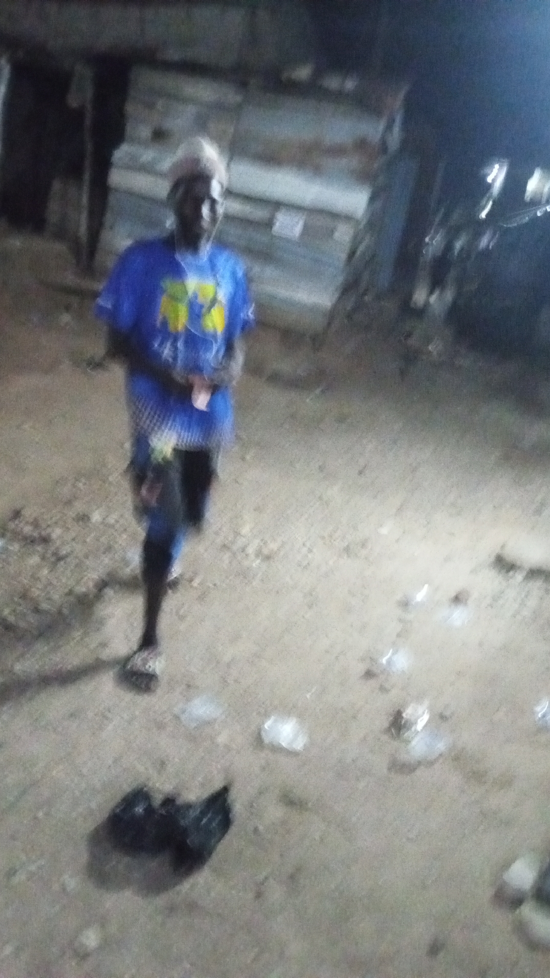 An individual wearing a blue shirt, dark pants, and light-colored headwear is walking on a dirt ground surface. Several small, light-colored objects and a black plastic bag are scattered on the ground. Behind the individual, there appears to be a makeshift structure constructed of metal sheets and wooden supports. The scene is poorly lit, suggesting it is either nighttime or a dimly lit area. Based on the provided location, this is likely a street scene in Adankolo, Nigeria.