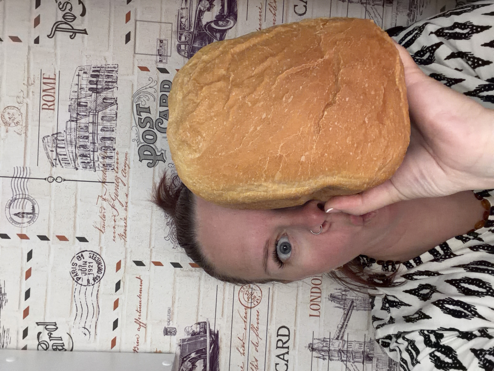 A person is playfully holding a fresh, golden-brown loaf of bread against their face, partially covering their nose and mouth, in a close-up indoor shot. The person, with fair skin, blue eyes, dark hair tied back, and a nose ring, looks directly at the camera with a wide-eyed, expressive gaze suggesting humor or surprise. They are wearing a black and white patterned top.
The loaf of bread appears freshly baked, with a slightly uneven, golden-brown crust.
The background is a light-colored surface, possibly wallpaper or fabric, featuring a vintage travel and postcard theme. Visible illustrations include line drawings of famous landmarks such as the Colosseum in Rome and Tower Bridge in London, along with an old car and various postmark designs. Text elements clearly visible on the background include "ROME," "LONDON," and "POST CARD" repeated multiple times. A specific postmark reads "PARIS 29 JUIN 1921 ORCHICEL," and French text "Bien affectueusement" (Affectionately yours) is also present. Other discernible words include "GOLDEN" and partial text such as "ard," "sune," "inkler," and "CA." The overall impression is lighthearted and casual, set in Chaikovsky, Russia.
