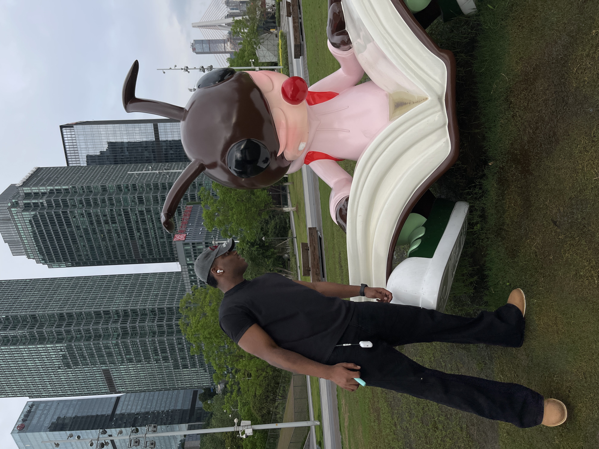 A man stands next to a large cartoonish sculpture in Jiangbei District, Chongqing, China. The sculpture features a brown-headed cartoon figure in a pink suit, with a red nose and a red bow, seemingly emerging from a white book with green-accented shoes. Tall modern buildings rise in the background, while trees and green spaces suggest an urban park setting. The man, wearing a black t-shirt, black pants, a gray baseball cap, and tan shoes, appears to be looking up at the sculpture. He is holding a blue object in his right hand, and a white object is visible on his pants. The weather appears overcast.