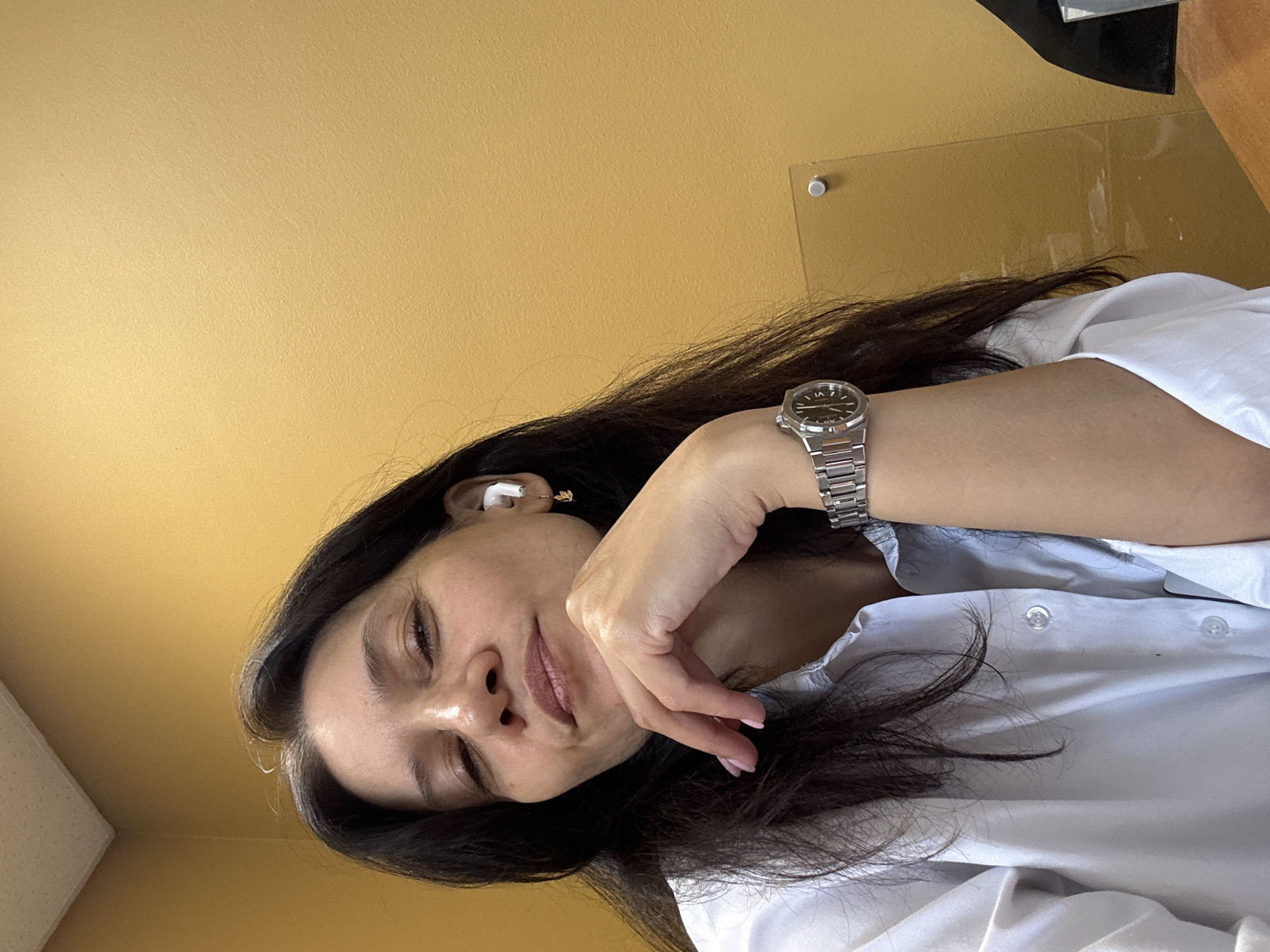 A woman with dark hair, wearing a white collared shirt, rests her chin on her hand. She is wearing white wireless earbuds and a silver wristwatch. The background is a mustard-yellow wall. A clear glass panel is visible on the right side of the frame. The image is a selfie, taken from a slightly high angle. There is no identifiable information suggesting the location is in the city of Chaikovsky, Russia.