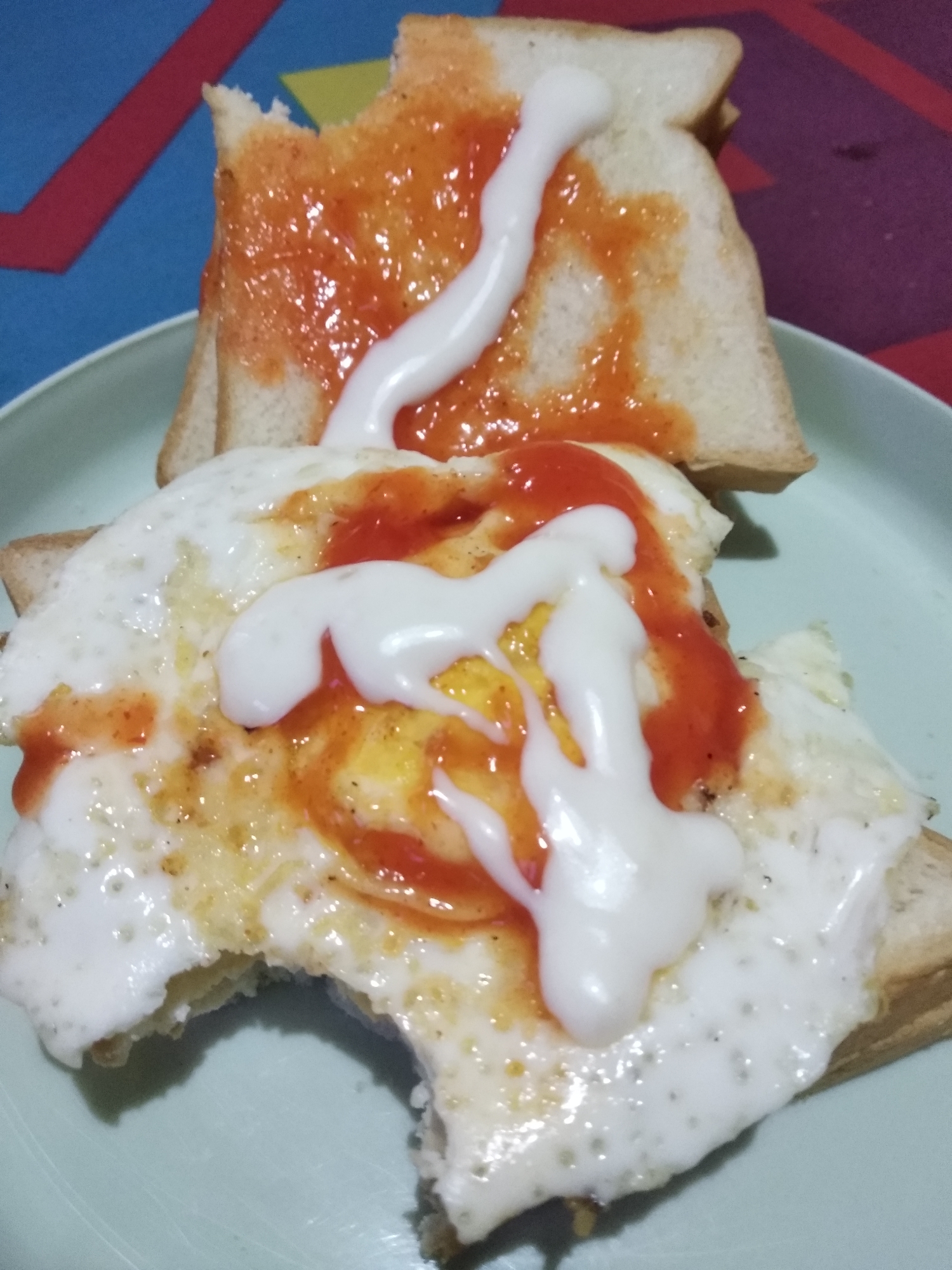 The image shows two slices of toast on a light blue plate. The bottom slice of toast is topped with a fried egg, ketchup, and mayonnaise. The egg appears to be sunny-side up with a runny yolk. The top slice of toast is topped with ketchup and mayonnaise. The background is a colorful, abstract pattern with red, yellow, and blue shapes. The setting is likely a home in Balaraja, Indonesia, given the style of the food and the commonality of such a meal in Indonesian households.