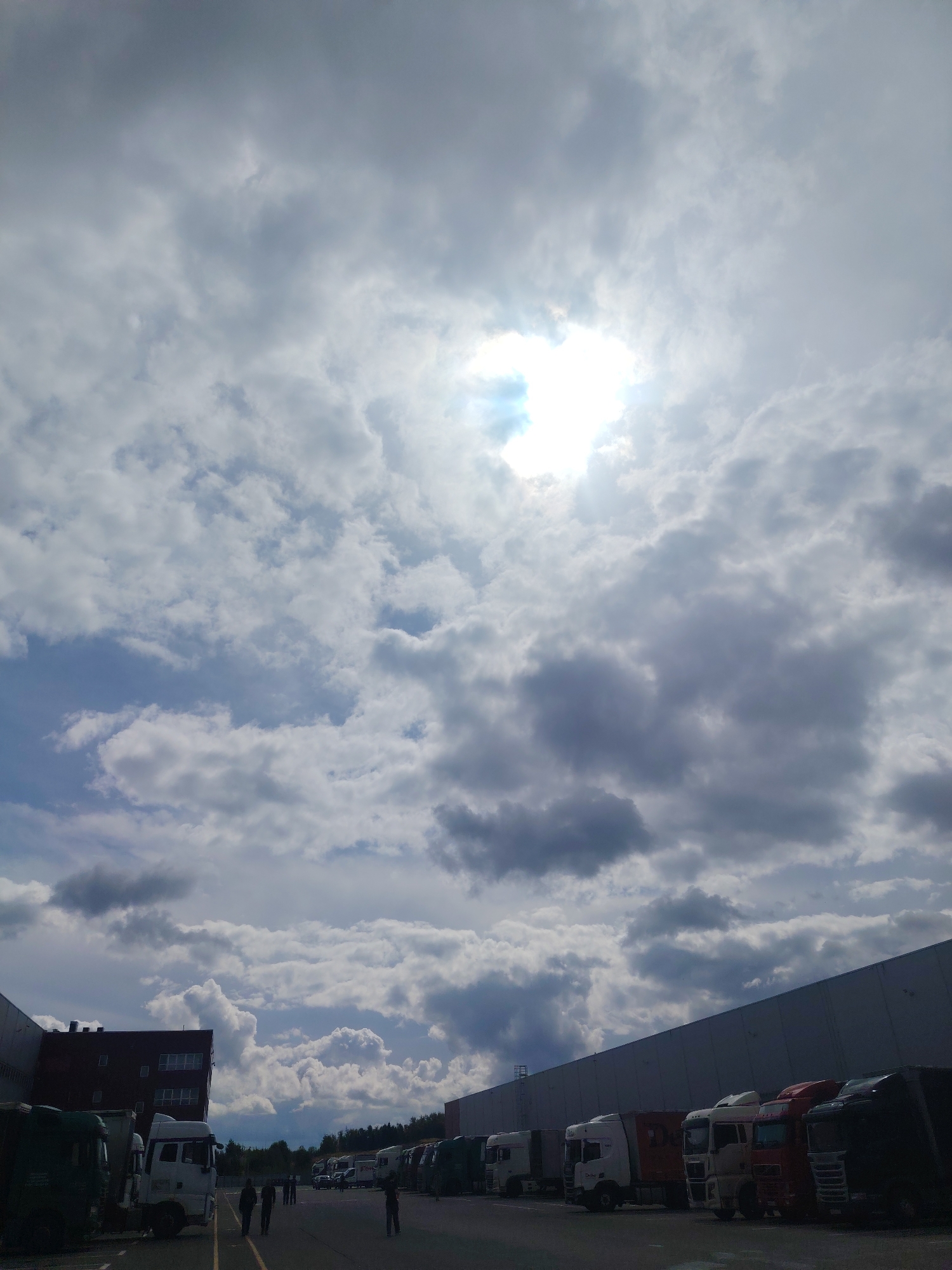 In the city of Хоругвино, Russia, a commercial area is depicted under a cloudy sky with the sun breaking through. The scene features a large, dark-colored building on the left, and a long, light-gray industrial building dominating the right side of the image. Several trucks, including semi-trailers and smaller transport vehicles, are parked in the lot between these structures. Some people are visible walking in the paved area. The ground is asphalt, with yellow markings indicating parking or traffic lanes. The overall impression is an industrial or logistics hub.