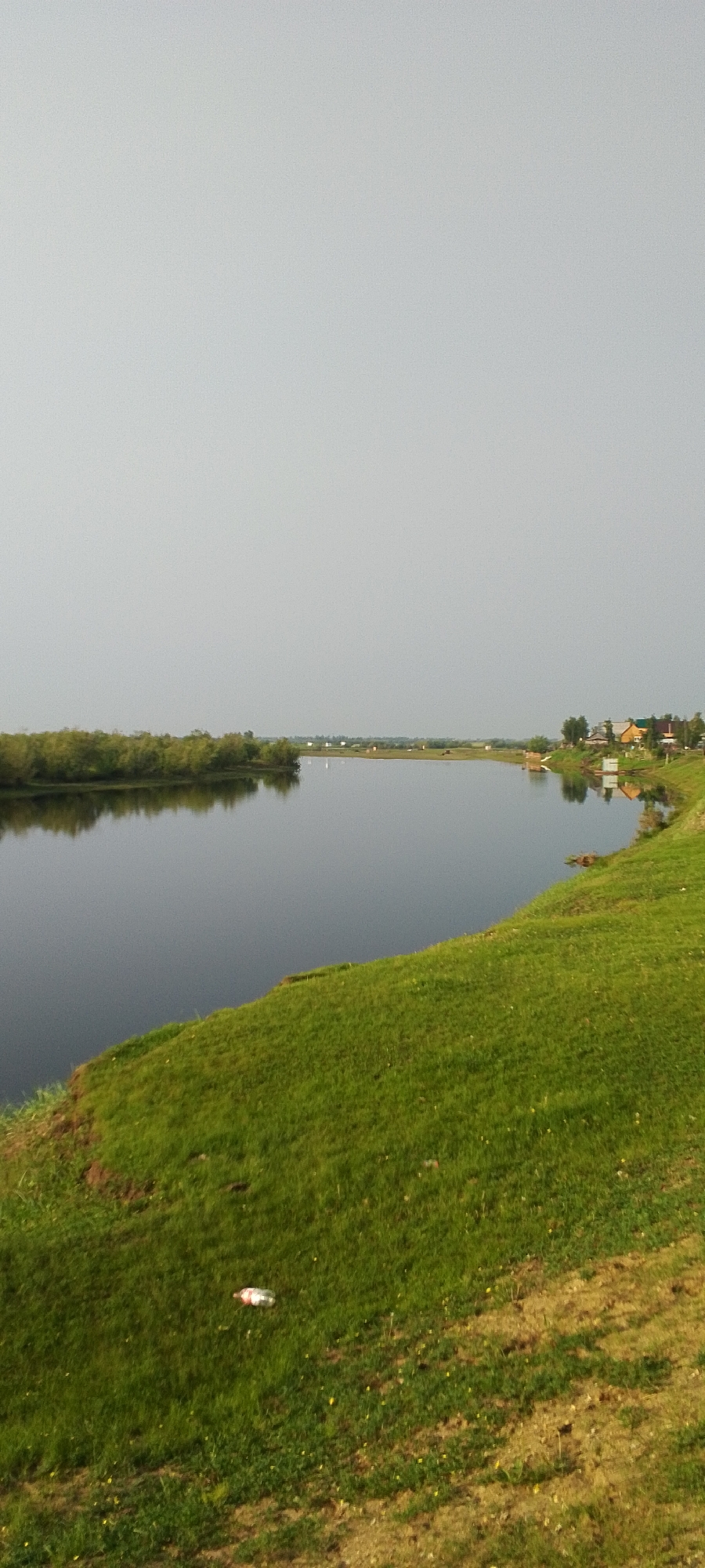 A serene landscape photograph captures a wide, calm river reflecting a pale, overcast sky. The riverbend is framed by lush green grass on the right bank, sloping upwards, and a dense line of trees on the left bank. In the distance, across the river, a small cluster of houses with red and yellow roofs dots the horizon, nestled amongst greenery. The overall scene is one of peaceful natural beauty, with a single plastic bottle lying on the grass in the foreground, a subtle sign of human presence in an otherwise tranquil environment. The location is the village of Tulagino, Russia.