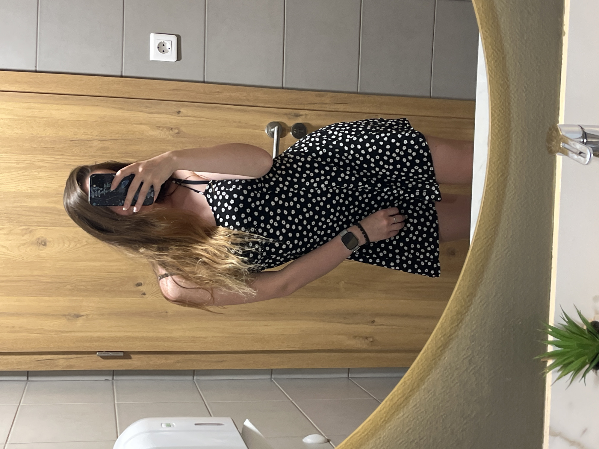 A young woman is taking a mirror selfie in what appears to be a public restroom, likely in Plovdiv, Bulgaria. She is wearing a short black and white polka dot dress and a smartwatch. The restroom has tiled walls, a wooden door with a silver handle, and a white soap dispenser. The mirror is large and reflects the woman, the door, and part of the sink area, which has a small artificial plant. The lighting is artificial and casts shadows.