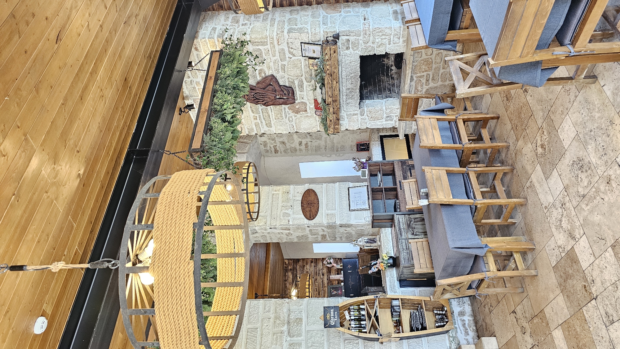 This image shows the interior of a restaurant in Kavarna, Bulgaria. The architectural style incorporates natural stone walls. A large, round chandelier made of rope and metal hangs from the ceiling. Tables and chairs with grey cushions are arranged on a stone-tiled floor. A fireplace is visible on one wall, adorned with decorative items and greenery. Other decorations include wood carvings, potted plants, and boat-shaped shelves filled with wine bottles. The overall ambiance is rustic and traditional.