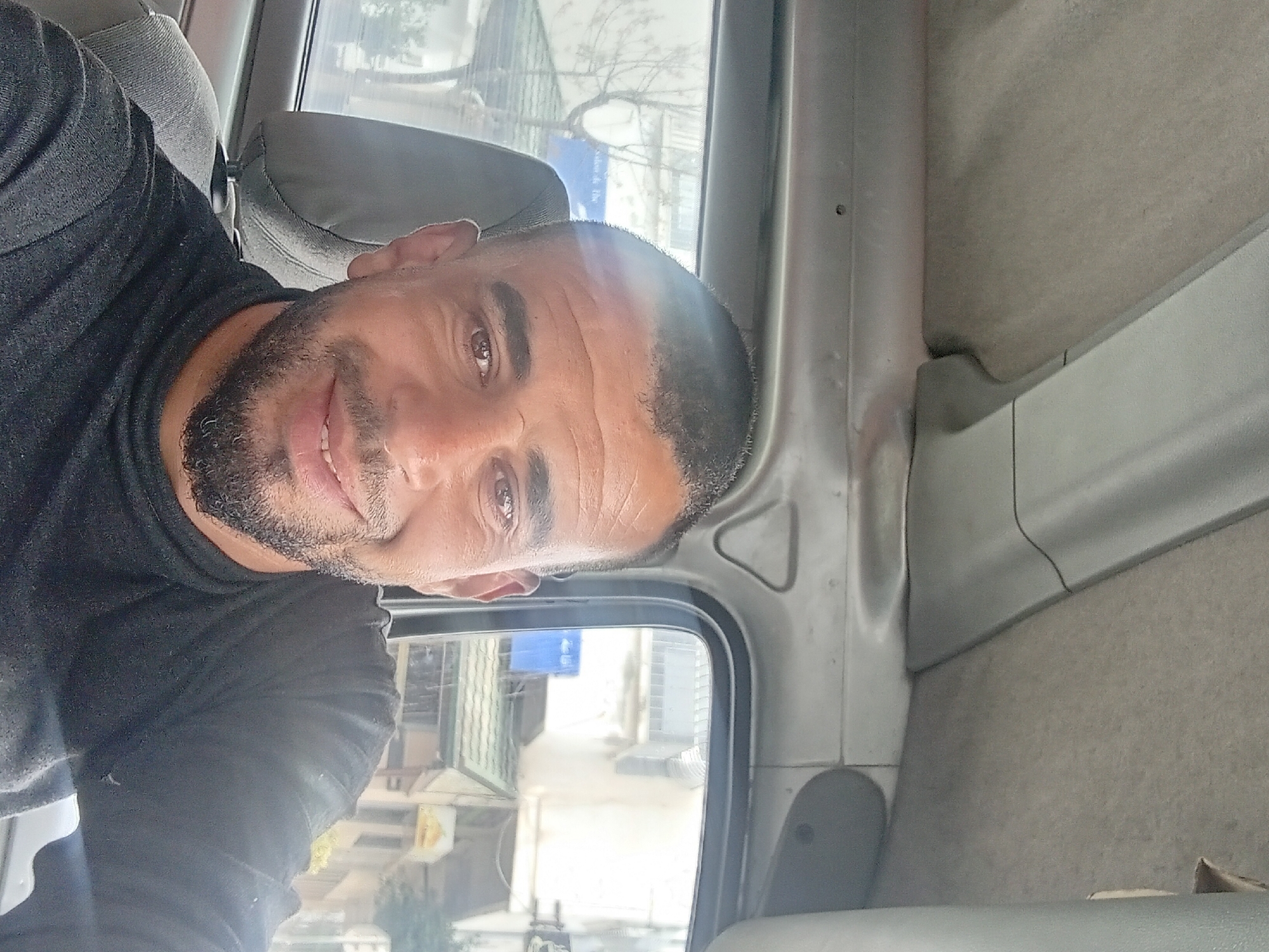 A selfie of a man with a beard in a black shirt taken inside a vehicle in Martil, Morocco, is rotated 90 degrees. Buildings and trees are visible through the vehicle's window. The interior of the vehicle is gray.