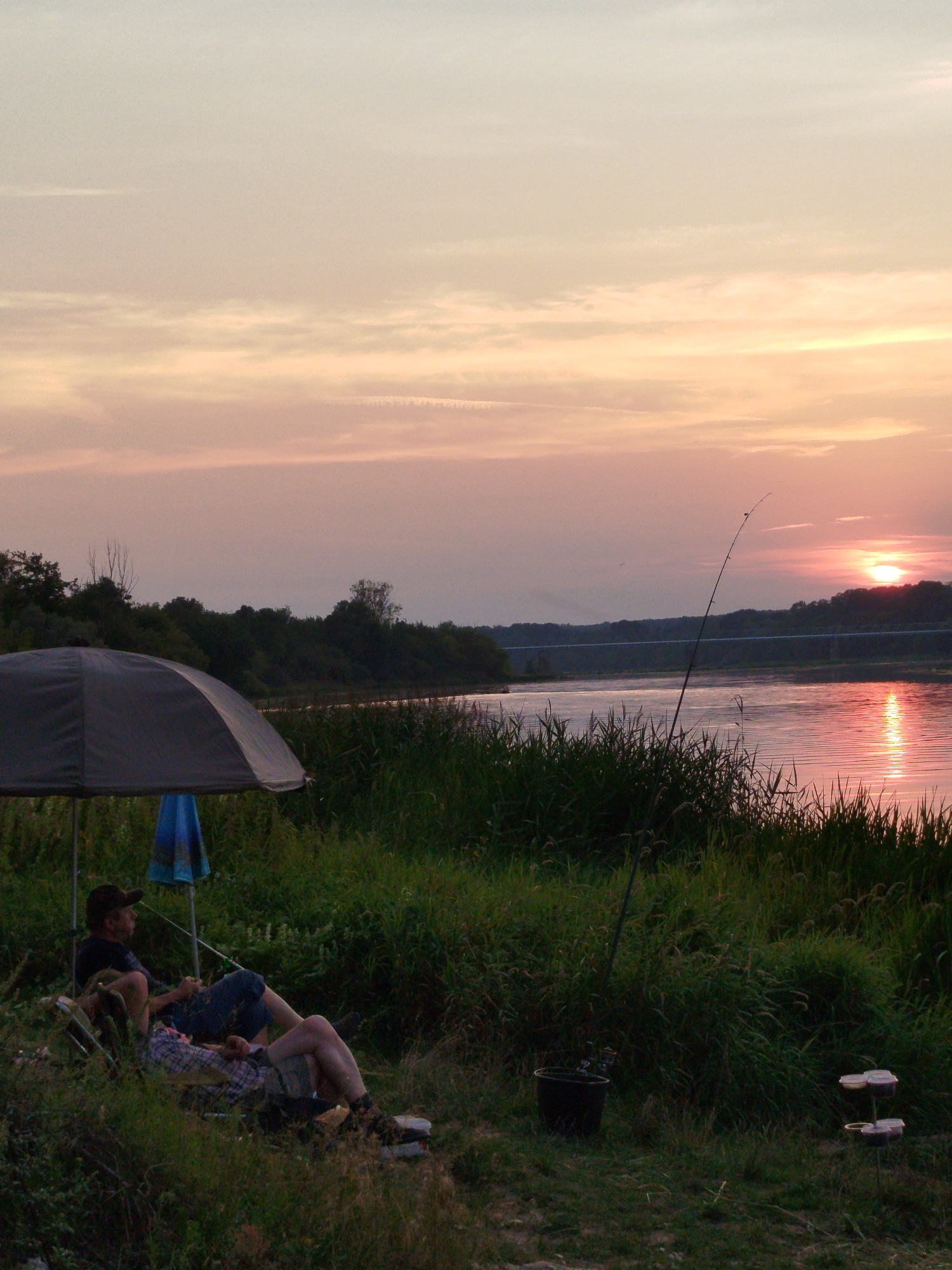 The image depicts a serene sunset scene by a river, likely in or near Topolina, Poland, given the context. Two individuals are relaxing in lawn chairs under a large dark umbrella. One person, wearing a dark cap and shirt, is seated with their legs extended. The other person, wearing a plaid shirt and shorts, is reclined, with their head resting on a cushion. Their interaction is not explicitly defined, but their relaxed postures suggest companionship. In the foreground, tall green grass and reeds line the riverbank. A fishing rod stands upright, leaning slightly, with its tip extending towards the sky. Further to the right, a black bucket and a small tiered stand with containers are visible, suggesting fishing equipment. The river reflects the warm hues of the sunset, with the sun setting on the horizon, casting a pinkish glow across the water. A bridge is faintly visible in the distance across the river, and a line of trees borders the opposite bank. The sky is a soft gradient of pale yellow, pink, and light purple, with wispy clouds. The overall atmosphere is one of peacefulness and quietude, characteristic of a summer evening by the water.