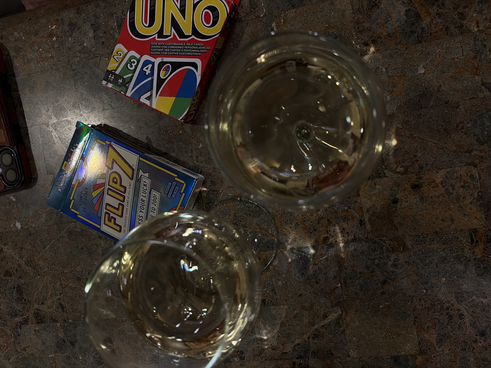The image captures a tabletop scene featuring several items. On the upper-left is a box of the card game "UNO", next to a box of "FLIPZ" card game, a cell phone with a dark brown case is at the left. Two wine glasses containing a clear liquid, possibly wine, are placed to the right of the card games. The surface appears to be a countertop, likely marble or a similar textured material. The photograph was taken indoors, probably in a private home. The image was taken in Trophy Club, United States.