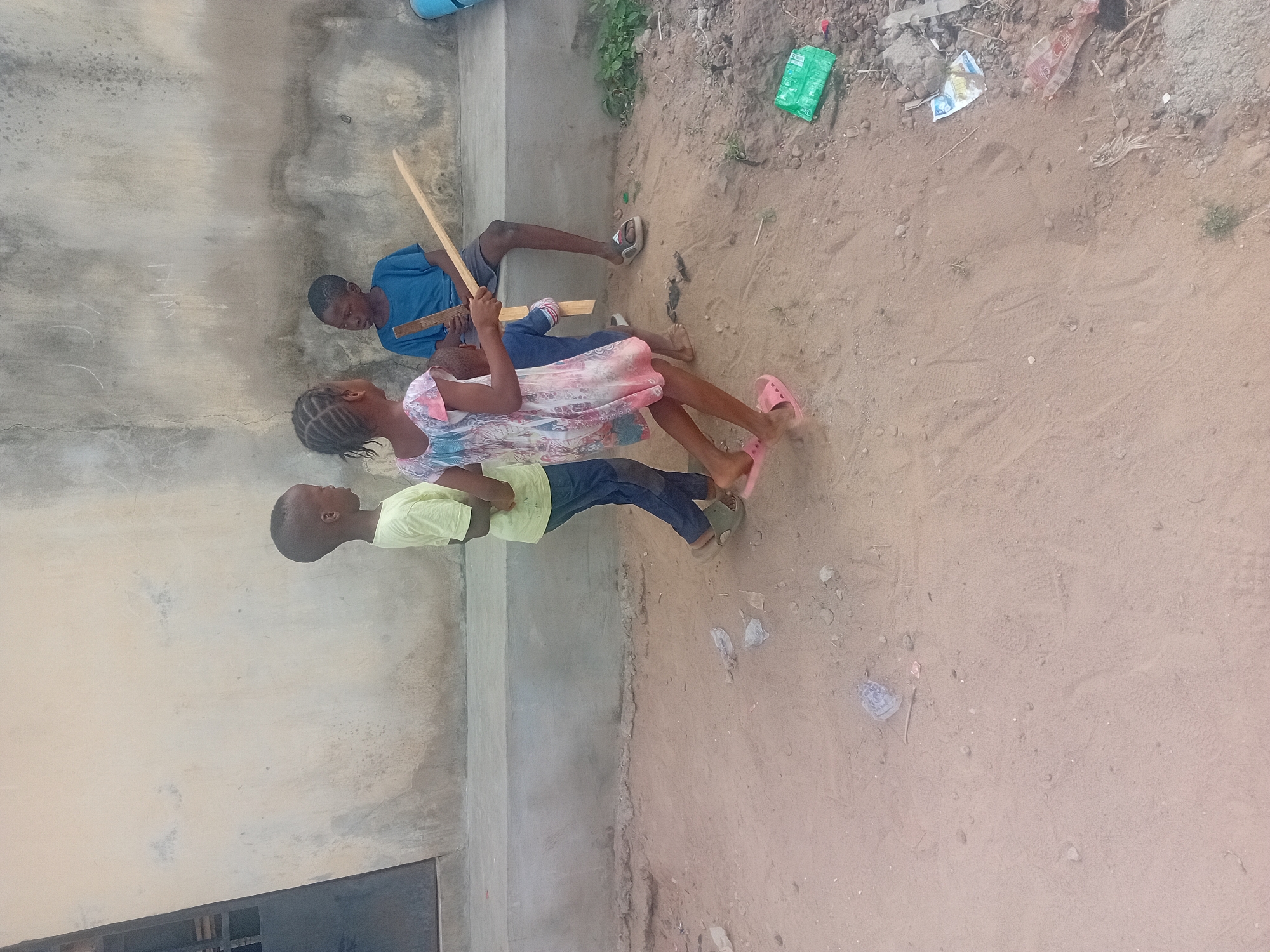 The image shows three children in an outdoor setting. A boy in a blue shirt is sitting on a ledge against a wall, holding two wooden objects. Next to him, a girl in a pink and white dress is lying on the ground. Below them, another boy in a yellow shirt and blue jeans is also lying on the ground. The ground is sandy with scattered debris, including plastic wrappers. The setting is adjacent to a concrete wall or structure. The location appears to be a simple outdoor area, likely in the city of Unknown, Unknown.