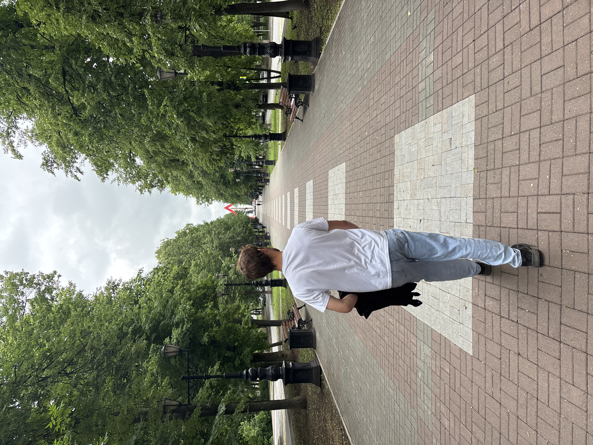 A young man with light brown hair, wearing a white t-shirt and light blue jeans, walks away from the camera down a wide, tree-lined pedestrian walkway. He is carrying a dark jacket over his left shoulder. The walkway is paved with reddish-brown bricks and features alternating square insets of lighter colored bricks. Ornate black lampposts line both sides of the walkway, interspersed with wooden benches and dark trash receptacles. Lush green trees with full canopies form a dense border on either side of the path. In the distance, at the end of the walkway, a tall, red and white structure, resembling a steeple or tower, is visible against a cloudy sky. The setting appears to be a park or public square in Ufa, Russia, likely in the vicinity of the Salavat Yulaev monument, given the architectural style of the distant structure and the overall park-like ambiance.