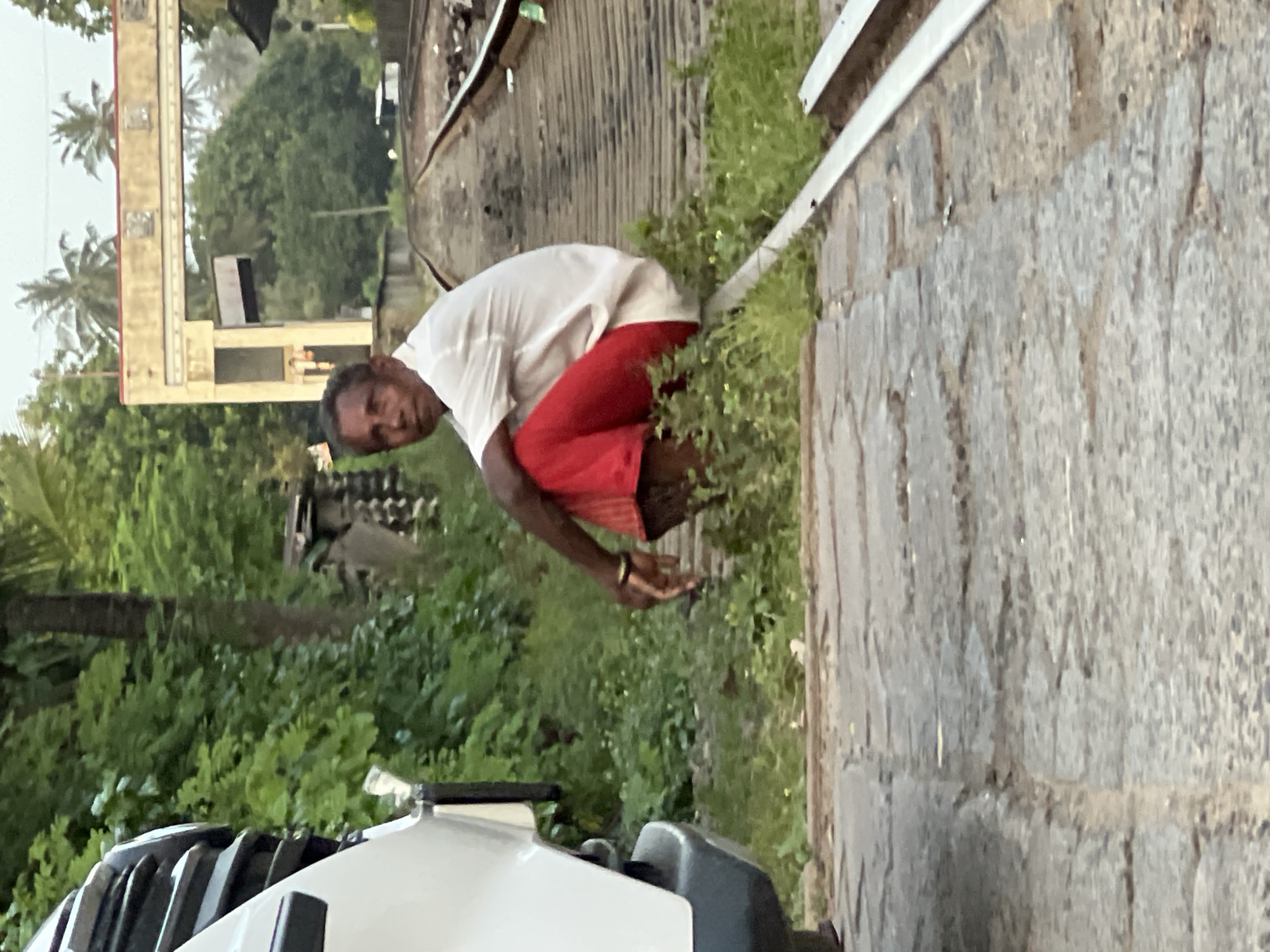 A man in a white shirt and red pants is sitting on a railroad track in an outdoor, rural setting. The man is looking towards the camera with a neutral expression. To the left of the frame, the side of a white vehicle is visible. The railroad tracks extend into the distance, with lush green vegetation on either side. In the background, a light-colored archway with some decorative elements can be seen, possibly part of a building or structure. The overall lighting suggests it is daytime, possibly late afternoon or early morning given the soft light. The ground in the foreground appears to be a mix of gravel and paving stones.