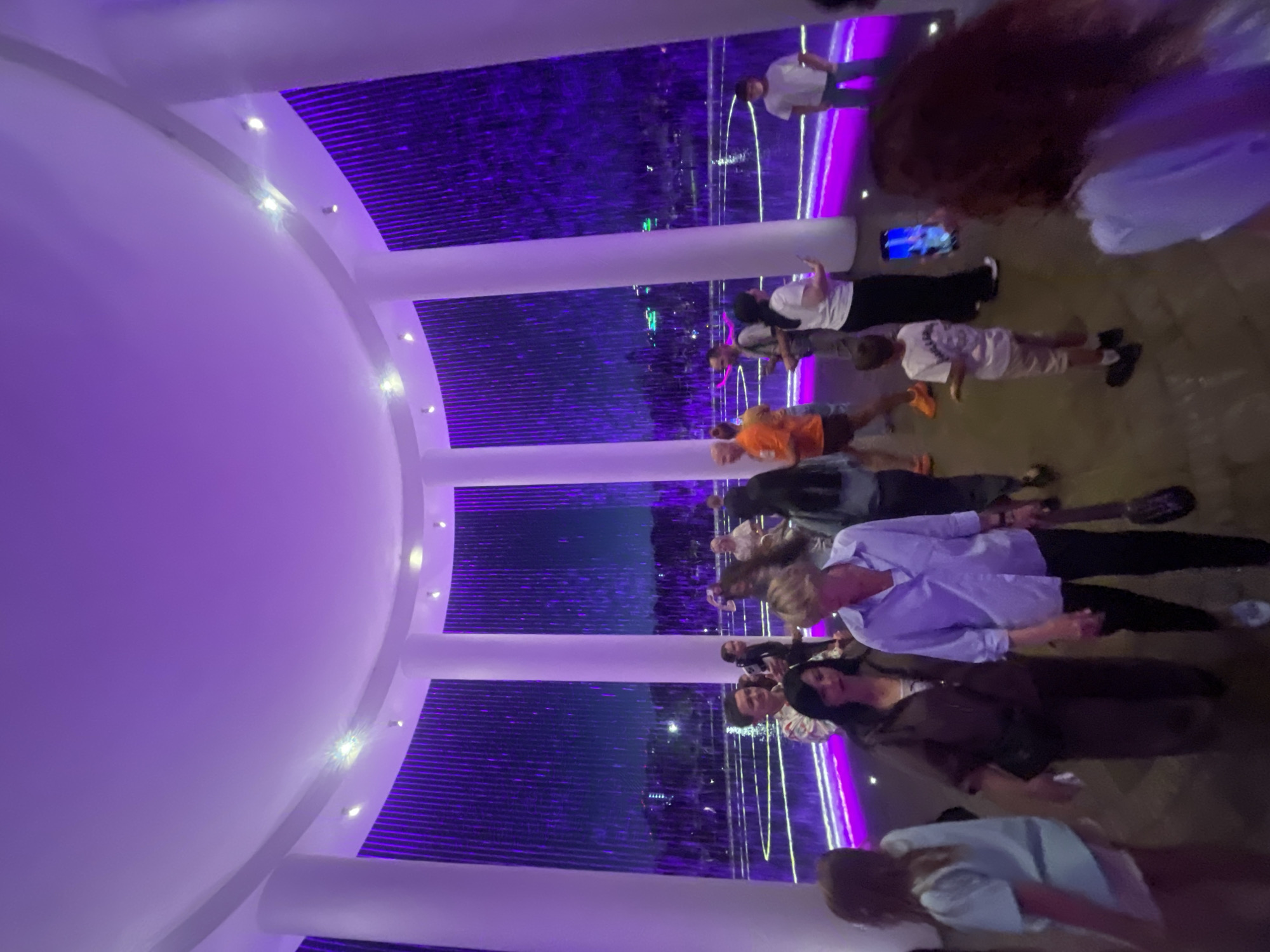 The image shows an indoor space with a circular white ceiling and white pillars. A purple, vertical light installation that resembles a waterfall of light is visible in the background. Several people, both adults and children, are present in the space, some of whom are looking at their phones or interacting with each other. The floor appears to be dark, with a circular, illuminated purple line near the base of the waterfall. Based on visual cues and common knowledge about public art installations, this scene is likely located at the "Fountain of Krasnodar" in Krasnodar, Russia.