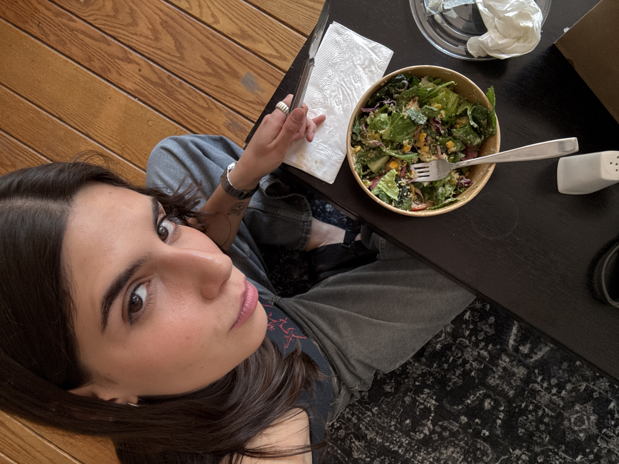 A woman with long, dark hair looks upward from a low angle, towards the camera. She sits with a table to her right that holds a salad in a round, light-brown bowl with a fork inside, a crumpled white napkin, and a salt shaker. Behind the bowl is a clear plastic container with crumpled white paper inside. The table is dark brown, and a portion of a black and white patterned rug is visible beneath it. The woman is wearing a gray top and gray jeans. A portion of wooden floor is visible to the left. It is impossible to determine the location in Mexico City based on the visual information provided.