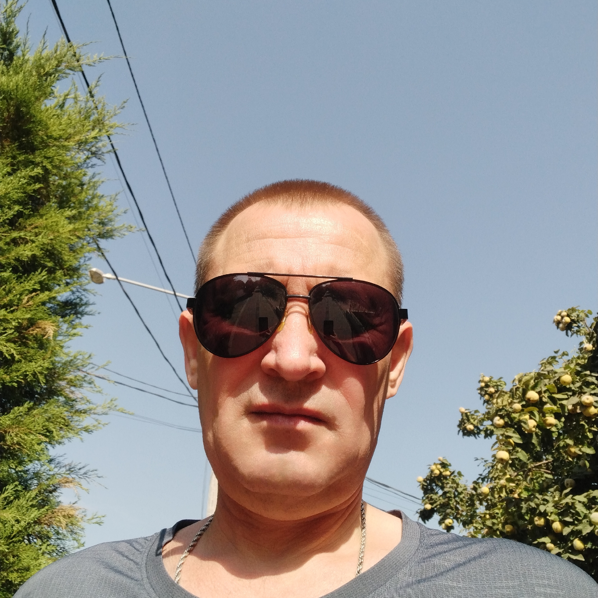 A close-up, low-angle selfie shows a fair-skinned man wearing dark aviator sunglasses and a grey t-shirt. He has short brown hair and appears to be outdoors under a clear blue sky, with green trees and power lines visible in the background. The man's face is centered in the frame, with the sunglasses reflecting a road and possibly a phone he is using to take the photo. A silver chain necklace is visible around his neck. The image quality is moderate with some visible graininess.