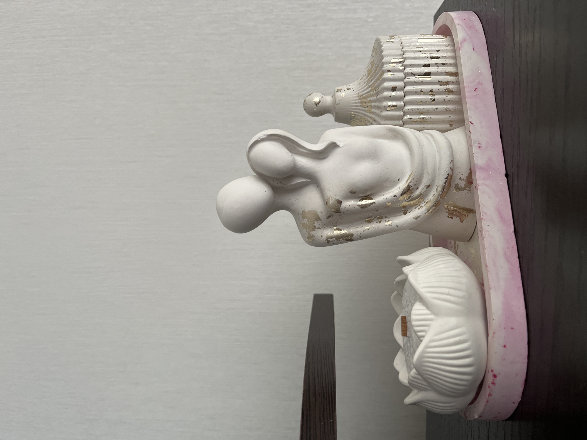 The image displays a decorative tray on a dark wooden surface. The tray is elongated and oval, with a pink and white marbled design. On the tray are three decorative objects. On the left is a white, lotus-shaped candle holder filled with white wax and a wooden wick. In the center is a white sculpture of a couple embracing, with gold flecks accentuating the figures. To the right of the couple sculpture is a small, white, lidded decorative box with a ribbed design and gold flecks. The background consists of a plain, light-colored textured wall and a dark horizontal shelf. There is no indication of the city of Ust'-Katav, Russia in the image.