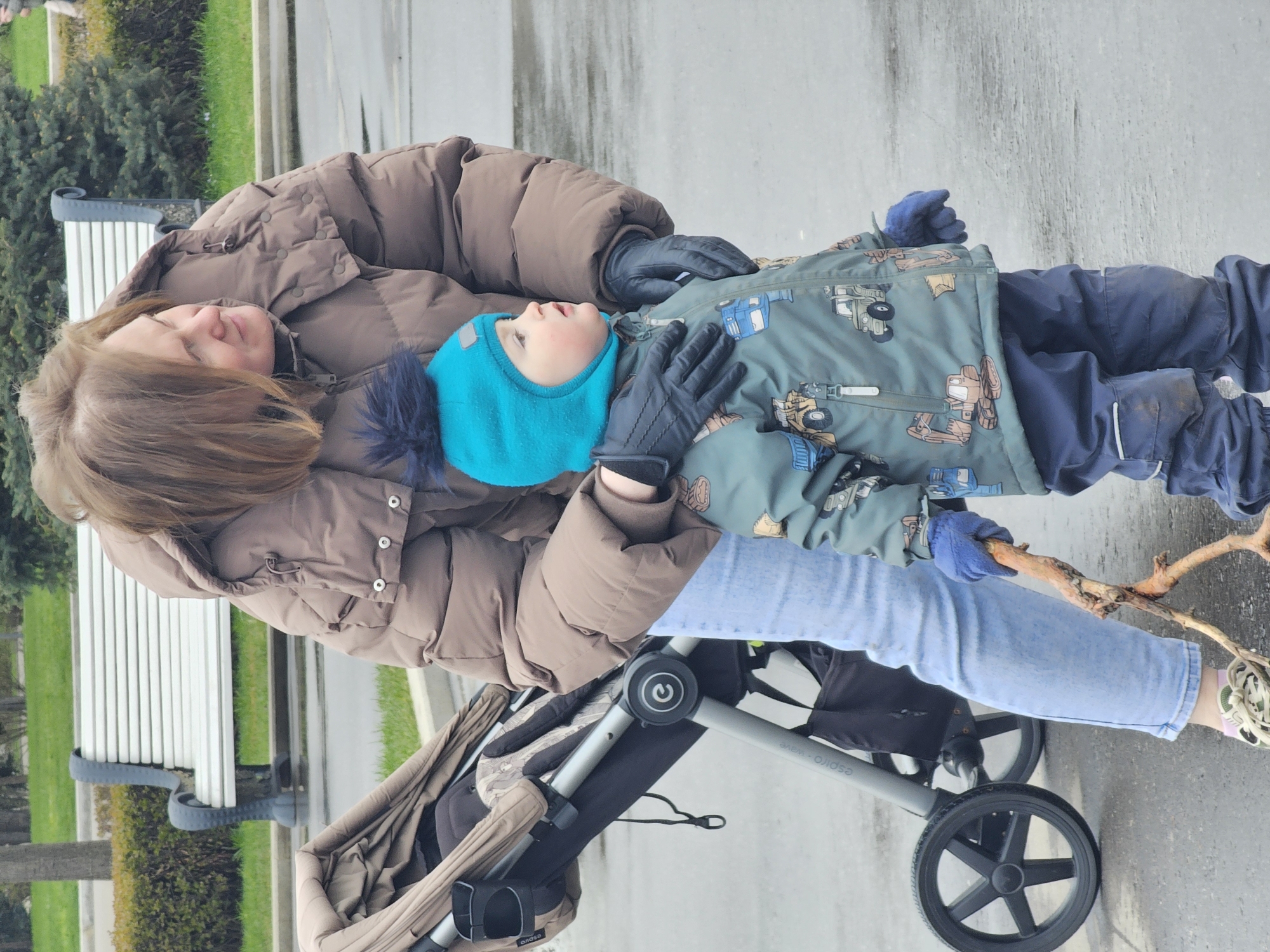 This vertical, full-shot image shows an adult and a young child outdoors, likely in a park or pedestrian area in Moscow, Russia. The ground is wet paved surface, suggesting recent rain or damp weather conditions.
The adult, a woman with long brown hair, is wearing a warm, brown puffer jacket, light blue jeans, and black gloves. She looks down at the child with a gentle expression, with her left arm around the child's shoulder.
The child, a toddler, wears a bright blue knitted hat, a green patterned jacket adorned with construction vehicles (like excavators and trucks), dark blue pants, blue gloves, and light sneakers with pink accents. The child looks upwards, holding a gnarled wooden stick in both gloved hands, seemingly engaged in play or observation.
In the background to the left, a white slatted park bench with dark supports is partially visible, along with green bushes and trees. A baby stroller, with a grey frame and black fabric, is visible in the lower left corner; the text "espra_wave" can be seen on its frame. The lighting suggests an overcast daytime.