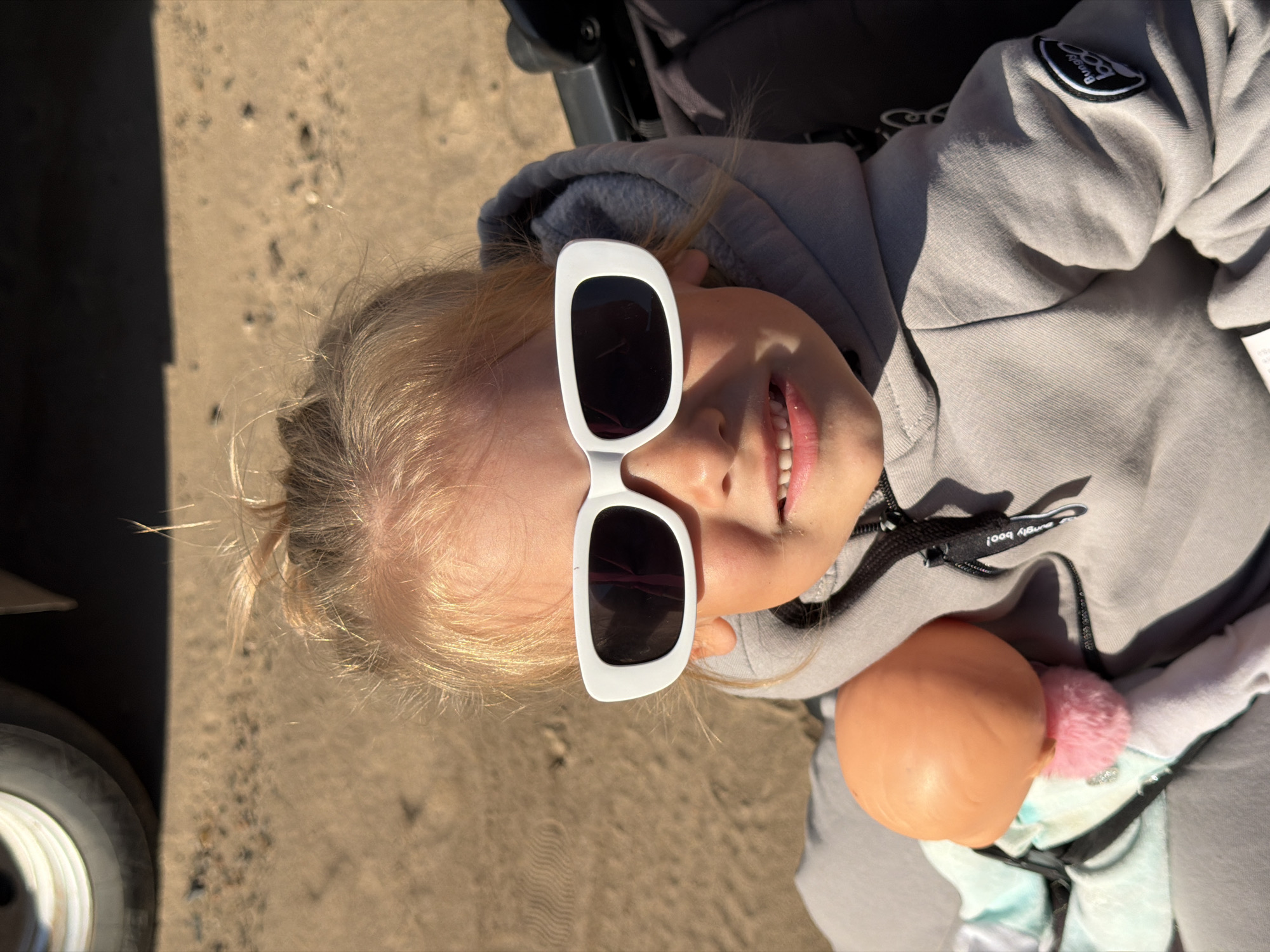 The image shows a close-up of a young girl wearing white rectangular sunglasses and a grey hooded sweatshirt. She is holding a doll with an orange head and a pink pom-pom. The background appears to be a sandy or dirt surface, possibly a beach or a playground, with a portion of a vehicle's tire visible in the upper left corner. The lighting suggests an outdoor setting on a sunny day. The girl is in a stroller, indicated by the stroller's fabric and handle. The scene is captured from a medium-close perspective, focusing on the girl's face and upper body. There is no information available to determine the specific location within Chaikovsky, Russia.