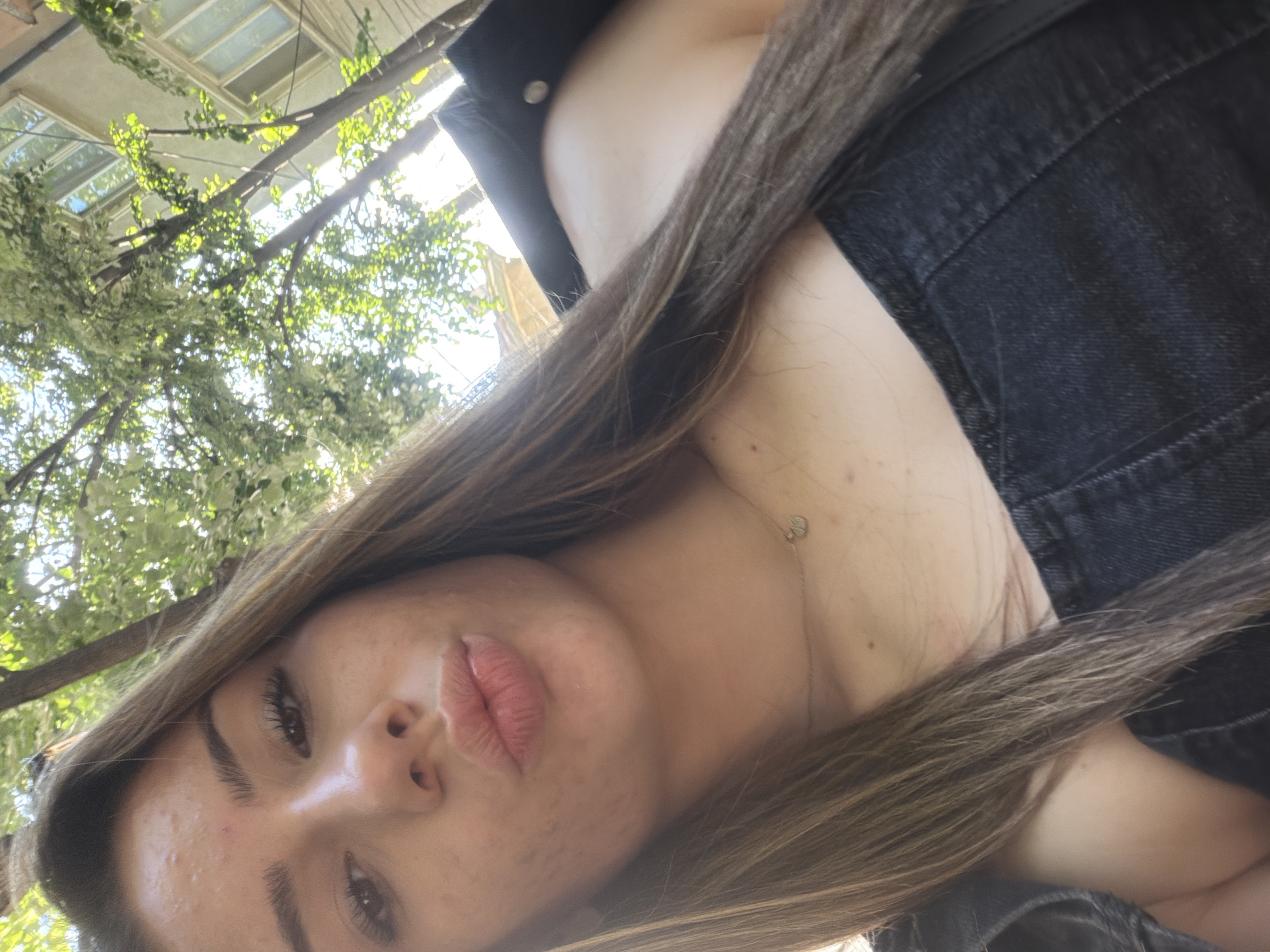 An outdoor, close-up, selfie-style photograph shows a young woman with long, brown hair and fair skin. She is wearing a dark denim top and a dark jacket. Her mouth is pursed as if she is blowing a kiss. A thin necklace with a small pendant is visible around her neck. In the background, sunlight filters through green leaves and branches of a tree, illuminating a building with windows. The scene is consistent with being in a city park or residential area in Shumen, Bulgaria.
