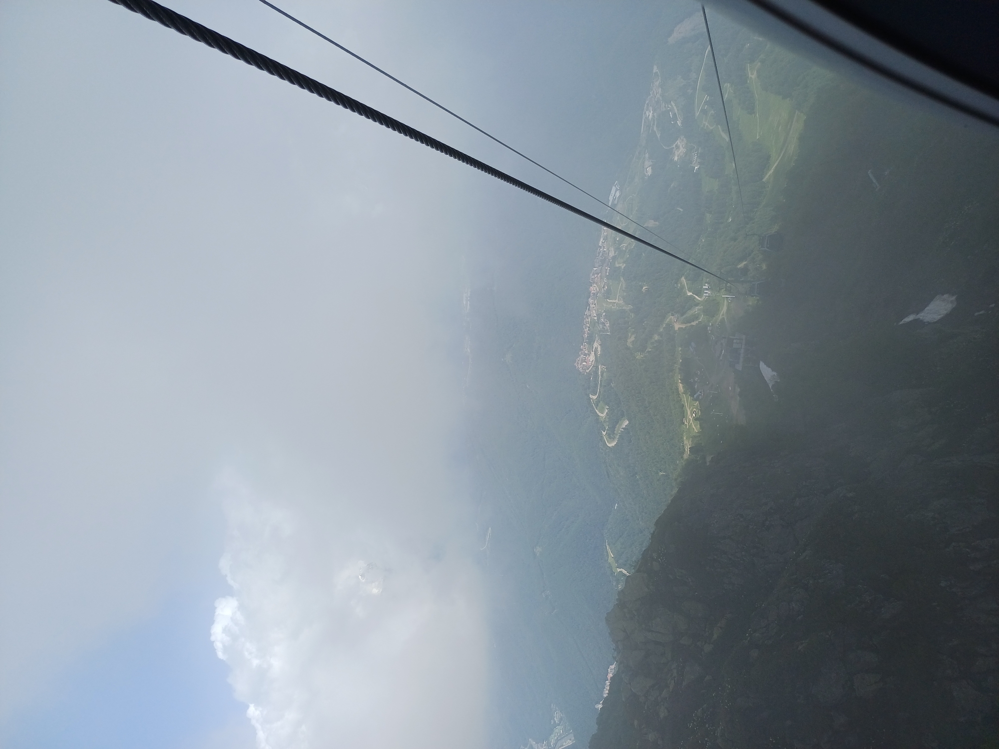 The image depicts a view from a cable car, looking down on a mountainous landscape. The cable car is evident, with thick black cables stretching across the frame. Below, a valley unfolds, revealing a network of roads winding through lush green forests. In the distance, a small town or village is visible, with buildings clustered together. The sky is partly cloudy, with patches of bright sky peeking through the overcast. The overall mood is one of awe and wonder, with a sense of scale and distance conveyed by the vastness of the landscape.
The image captures a moment of travel, likely a scenic journey or an ascent to a viewpoint. The time of day appears to be daytime, judging by the light. The weather seems mild, with a mix of sun and clouds. The location is identified as Estosadok, Russia, which is known for its mountain resorts and natural beauty. The presence of the cable car suggests a tourist or recreational setting. The image is dominated by natural elements, with the man-made structures of the town and roads providing a contrast to the wilderness. The photograph is devoid of any visible text.
