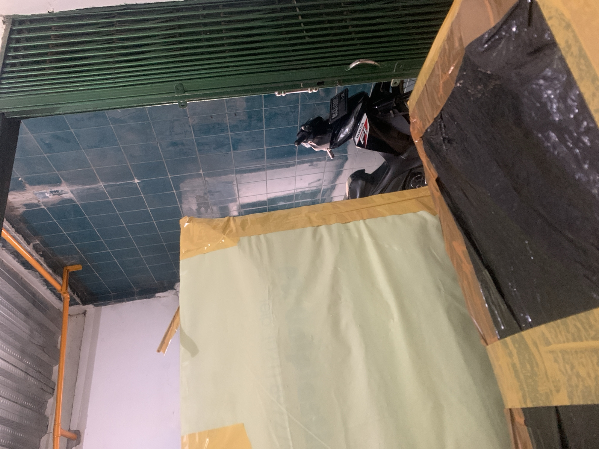 The image shows an indoor scene with tiled walls and a metal ceiling. In the foreground, there are two large packages wrapped in plastic and tape. The larger package is covered in a pale yellow material with yellow tape, and the smaller package underneath is wrapped in black plastic with yellow tape. To the right, there is a green metal gate, partially open, revealing a dark interior. A black motorcycle with a license plate is parked inside the opening, with the handlebars and part of the seat visible. The license plate appears to be from Indonesia. The overall impression is of a storage or workshop area. Without further information or context, it is not possible to pinpoint the exact location in Palembang, Indonesia, but the presence of the Indonesian license plate and the general architectural style suggest it is within Indonesia.