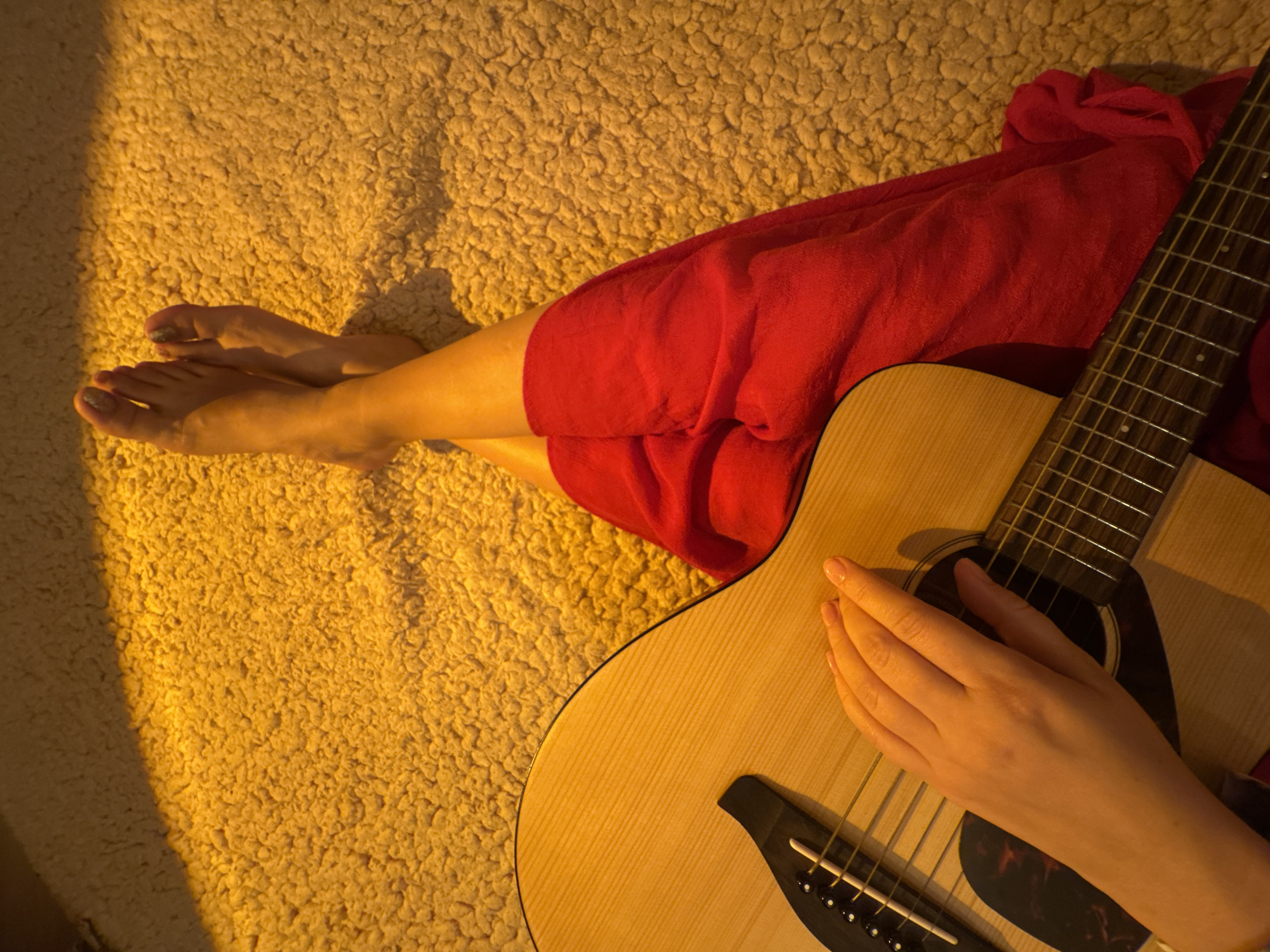 A top-down, close-up shot shows a person's hands and feet on a yellow, shaggy carpet. The person is wearing a red dress and is holding an acoustic guitar. The lighting is warm and casts shadows on the carpet. The scene is indoors, but the specific location in the city is unknown.