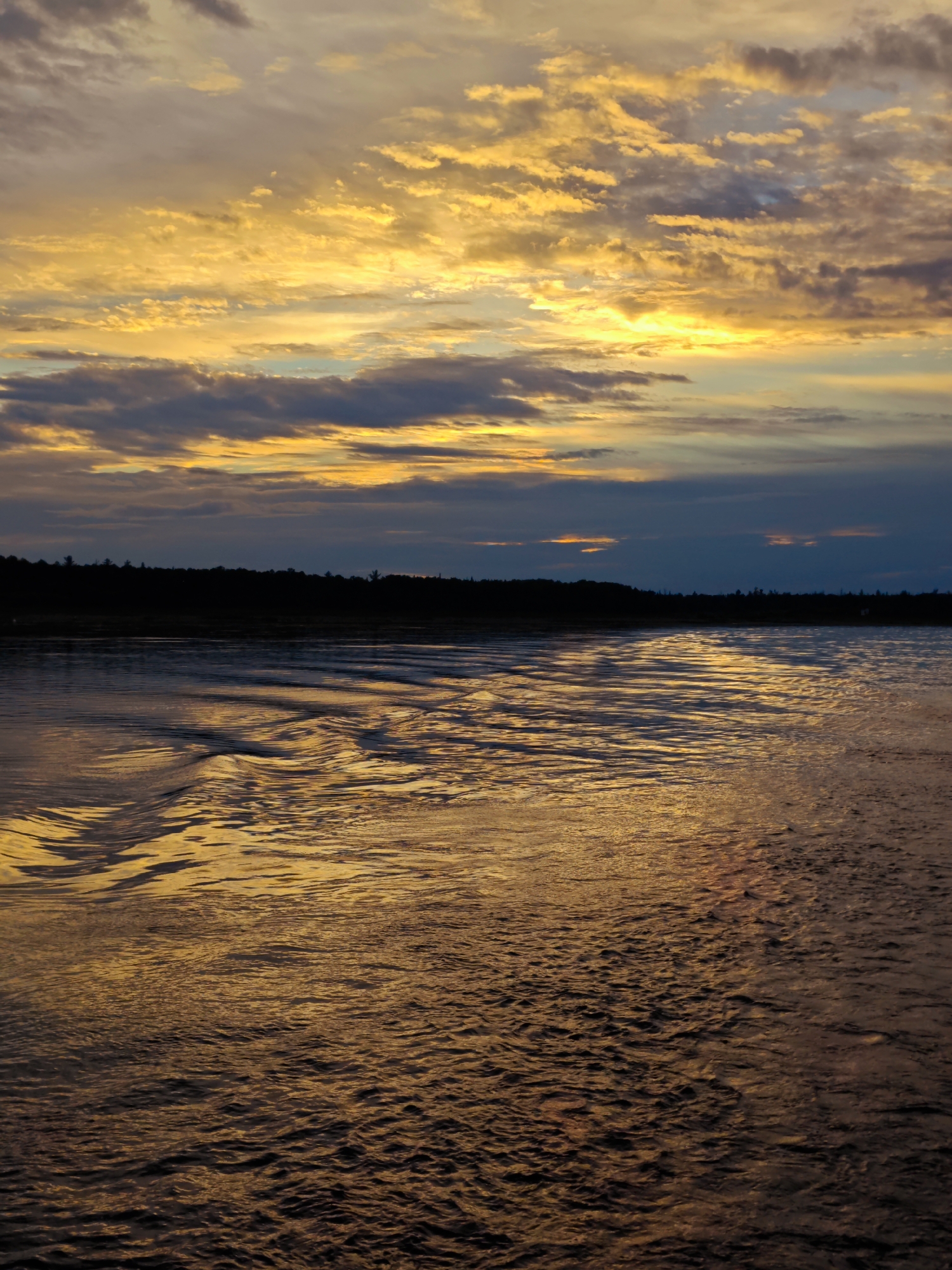The image depicts a serene natural landscape with a focus on water and sky. In the foreground, a body of water exhibits subtle waves, reflecting the warm hues of the sunset. The water's surface is textured by ripples, creating a dynamic interplay of light and shadow. In the middle ground, a dark, silhouetted treeline forms a distinct horizon, separating the water from the sky. The sky above is a canvas of dramatic sunset colors, with vibrant oranges, yellows, and softer blues and grays blending together. Clouds are scattered across the sky, catching the last rays of sunlight and adding depth and texture to the celestial scene. The overall impression is one of tranquility and natural beauty. There are no identifiable structures, people, or activities visible in the image.