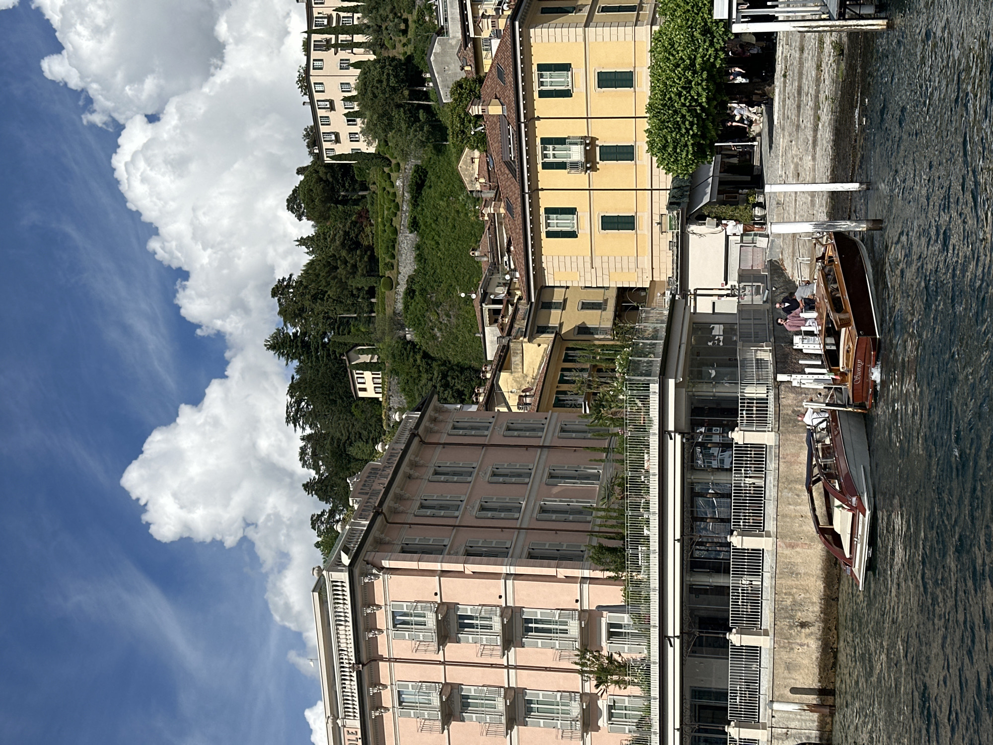 This image captures a vibrant lakeside scene in San Giovanni, Italy, under a bright blue sky dotted with large white cumulus clouds. The foreground consists of dark blue-green water, showing ripples and movement. Two classic wooden boats are moored near a stone or concrete embankment on the left. The larger boat, closer to the left, is dark reddish-brown with a light-colored stern cushion and a clear windshield. A smaller, darker brown wooden boat, possibly labeled "Sunny" on its stern, is positioned further right.
Above the water, a white-railed promenade or patio area features tables and chairs, suggesting an outdoor dining space, located beneath a multi-story, light pink building on the left. This building, likely a hotel given the partial "ALBERGO" sign visible on its roofline, exhibits classical European architecture with numerous windows, each fitted with light grey louvered shutters and small, ornate iron balconies. Balustrades adorn its roofline and upper facade.
To the right, a prominent multi-story yellow building with a red-tiled roof stands. It also features classical elements, including windows with dark green louvered shutters and a small white balcony on its upper floor. Several individuals are visible on the stone steps and along the shoreline near the boats, some standing by the mooring posts. Further inland, a lush green hillside rises steeply in the background, densely covered with trees and featuring terraced gardens with manicured hedges. Several additional light-colored buildings with dark shutters are visible nestled among the trees on the upper slopes of the hillside.