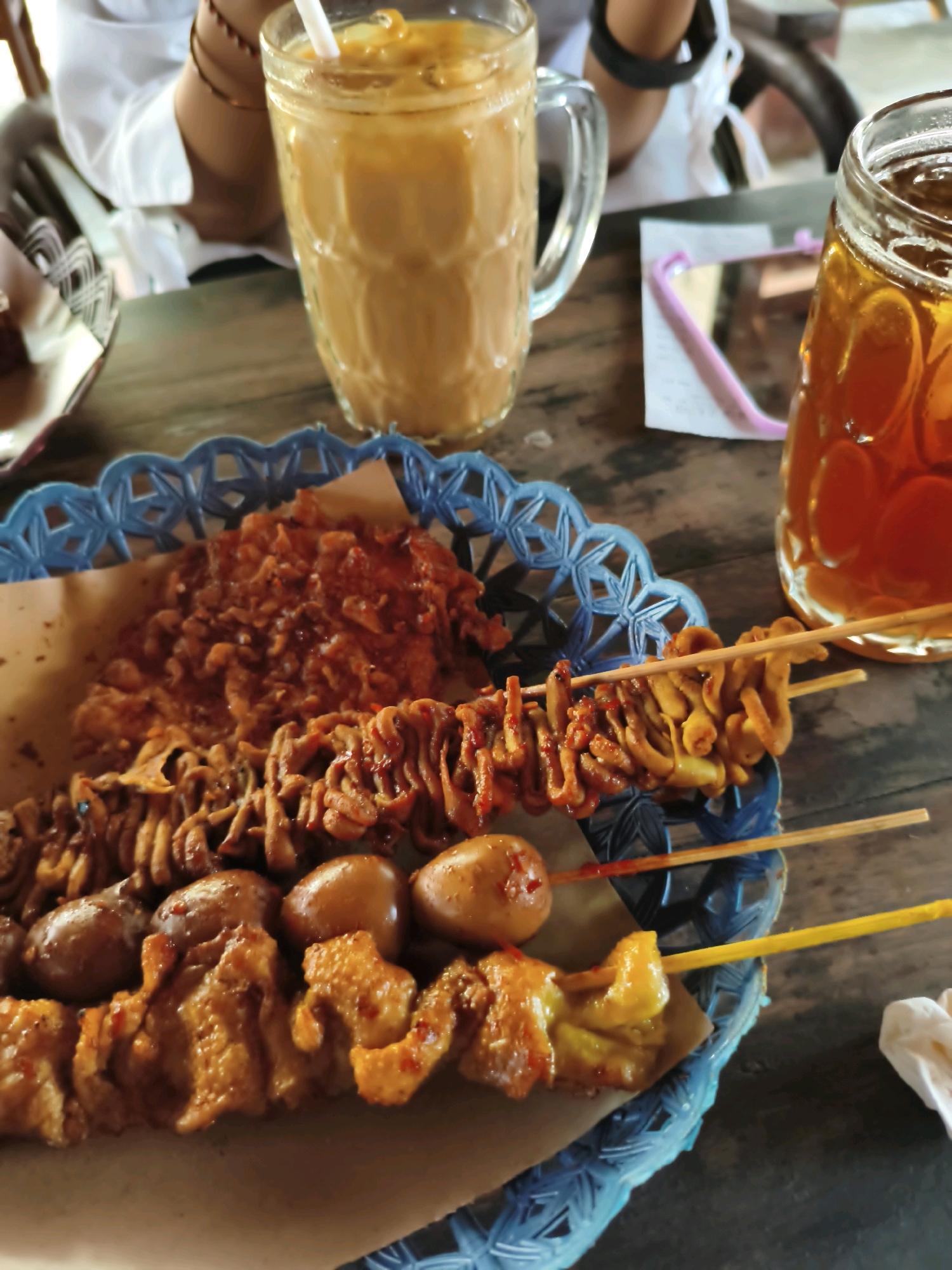 The image shows a close-up of a blue woven basket containing various fried snacks on a wooden table. The snacks include what appear to be fried chicken pieces, quail eggs, and intestines on skewers, all coated in a reddish sauce. To the right of the basket are two glasses, one filled with a light brown, frothy beverage (likely iced coffee or tea with milk) and another with a dark amber liquid (likely iced tea). In the background, a person is visible wearing a white shirt, with their arms and hands blurred. There is also a pink object and a white paper visible on the table. The lighting suggests an outdoor or semi-outdoor setting. The overall scene depicts a casual dining or snack experience.