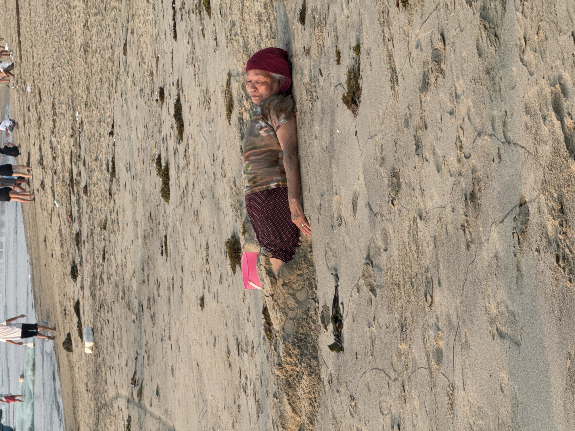 An older woman with a red head covering, patterned shirt, and polka-dot pants lies on a sandy beach. A pink bucket sits near her legs. The beach stretches out, with other people visible in the distance near the water's edge. The beach appears to be in Đà Nẵng, Vietnam.