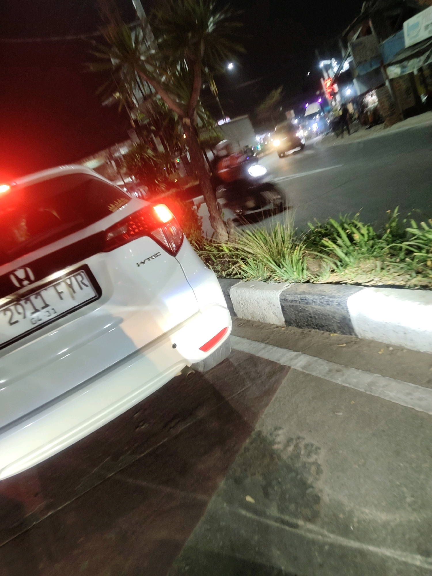The image shows a white Honda HR-V parked on the side of a street at night, likely in Mekarmukti, Indonesia. The car's rear taillights are illuminated. A license plate that reads "2911 FYR" is visible, along with "04-31" below it. The car is positioned next to a curb that is painted with black and white stripes. The street is paved and appears wet in places, possibly from recent rain. On the right side of the image, there is a patch of green vegetation planted between the curb and a sidewalk or road. In the background, other vehicles and buildings can be seen, illuminated by streetlights and the headlights of cars, suggesting an urban environment. The overall scene depicts a typical street at night with parked vehicles and passing traffic.