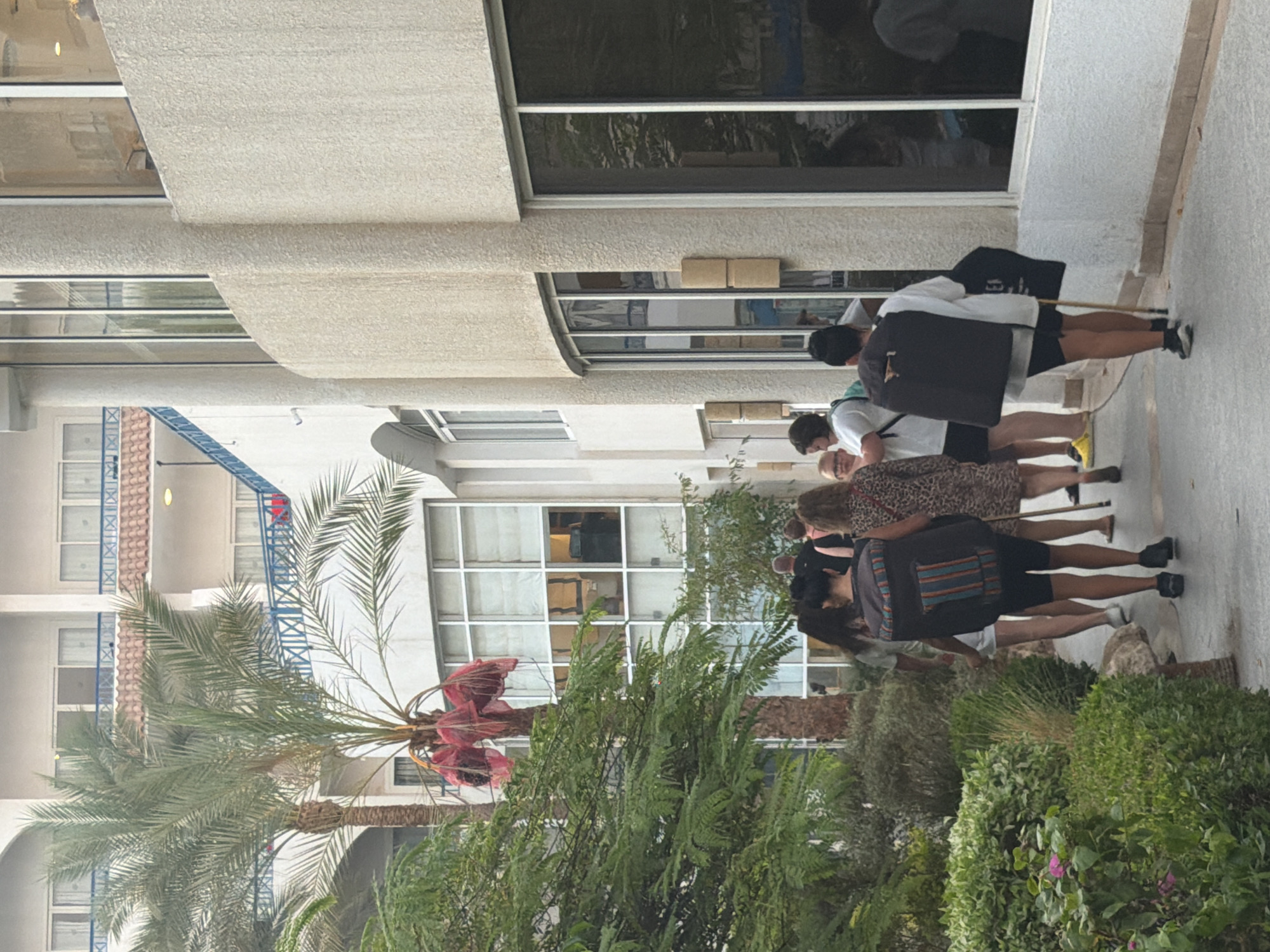 The image depicts a group of people walking along a paved pathway in front of a resort-style building. The building has a white, textured exterior and features large windows, some of which reflect trees and the sky. There are palm trees and lush green vegetation flanking the pathway and the building. The people are dressed in casual attire, and some are carrying bags. One person in the foreground is wearing a large backpack. In the background, a multi-story building with balconies and blue railings is visible, characteristic of hotels or resorts in a location like Sharm El Sheikh, Egypt, which is known for its coastal resorts and warm climate. The lighting suggests it is daytime, possibly late afternoon or early morning given the soft light.