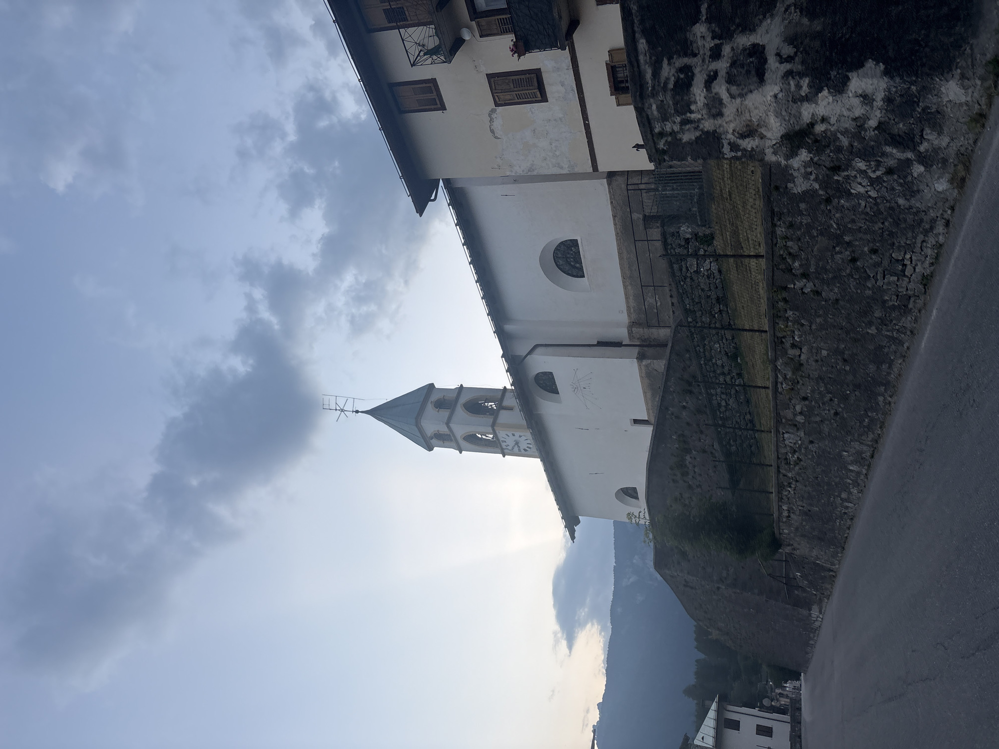 The image shows a church with a clock tower in an Italian village. The church is white with a green-roofed spire. A clock face is visible on the tower. Adjacent to the church is a building with shutters and balconies. The buildings are situated on a slope with a stone wall. A road runs in front of the buildings. The sky is overcast with some patches of sunlight breaking through. The overall scene suggests a rural European setting.